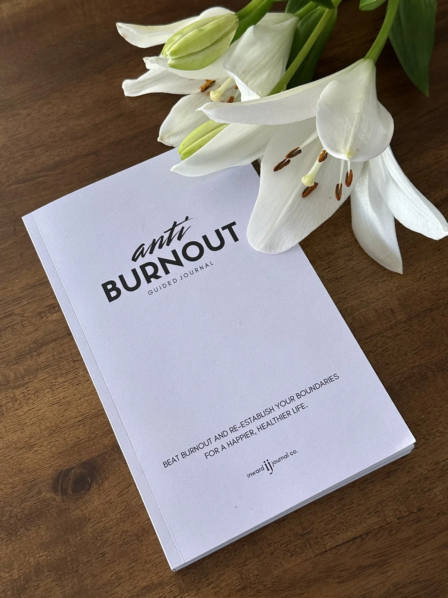 A white guided journal titled 'Anti Burnout' placed on a wooden surface, partly covered by white lilies.