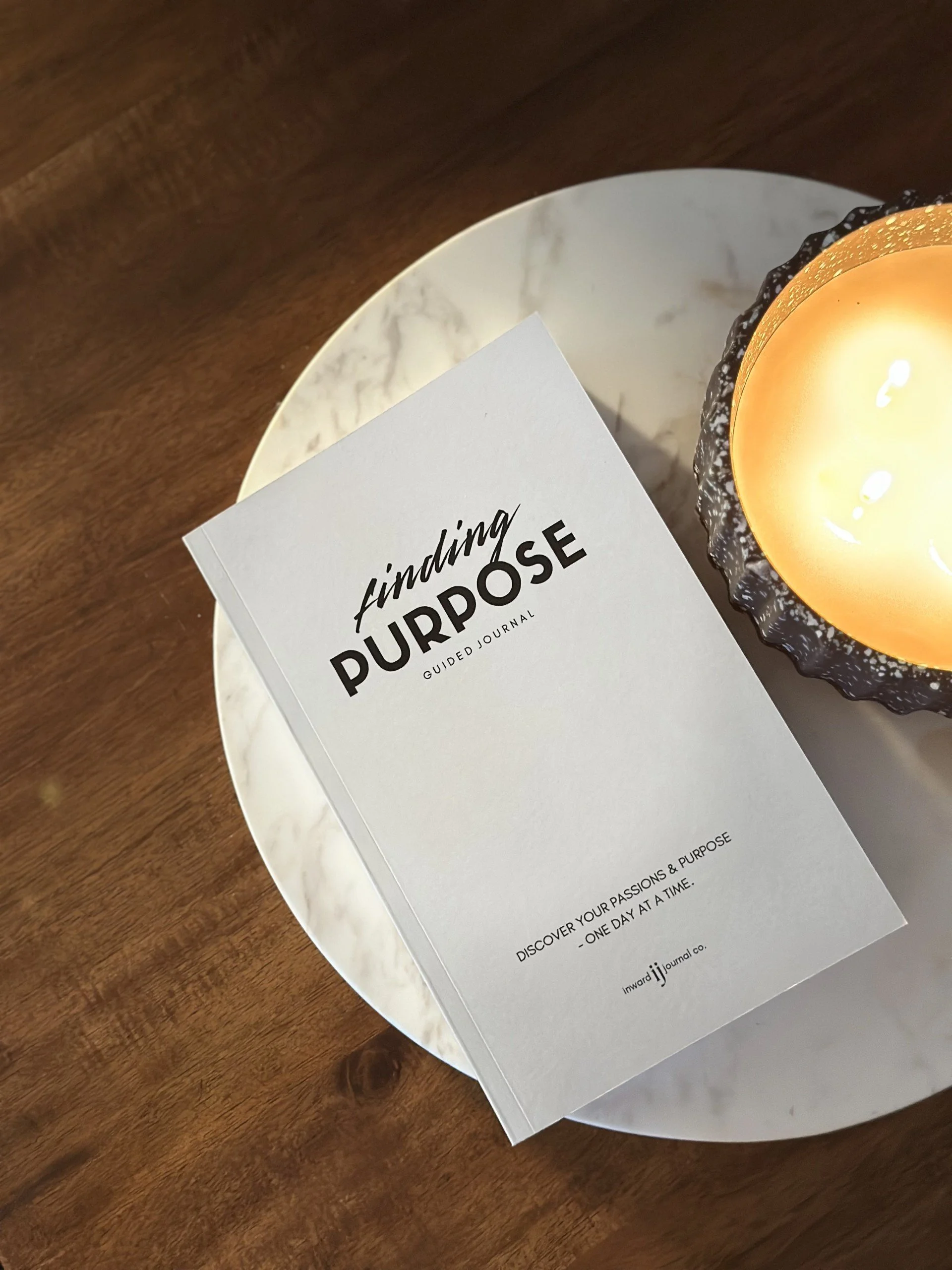 A guided journal titled 'Finding Purpose' on a round marble tray next to a lit candle on a wooden surface.