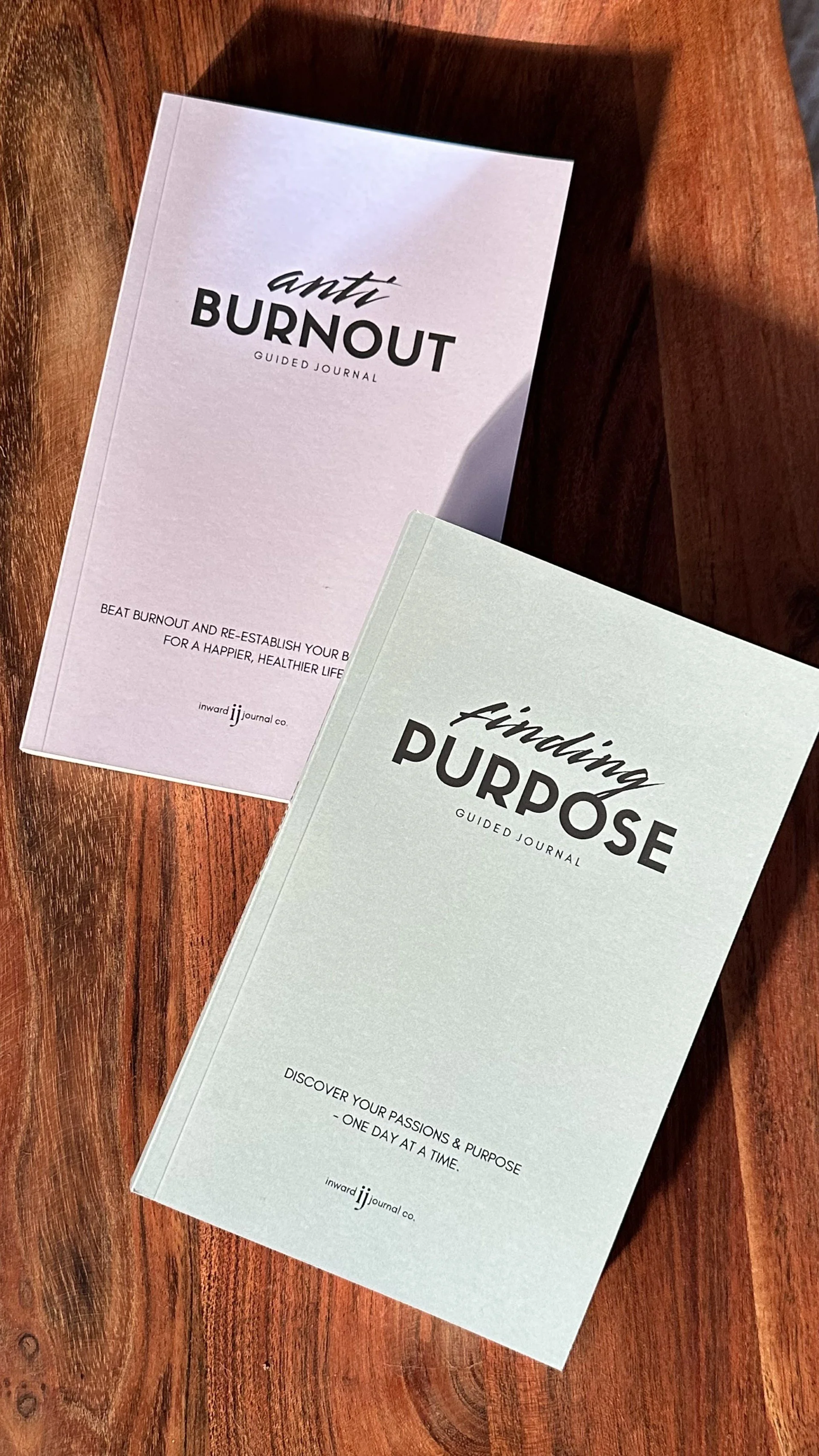 Two guided journals, one titled 'ant BURNOUT' and the other titled 'Finding PURPOSE,' placed on a wooden surface.