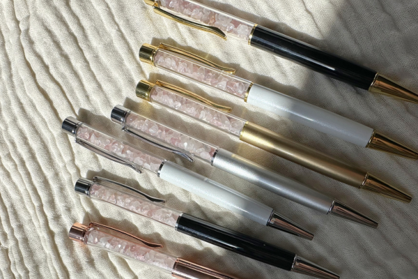 Six decorative ballpoint pens with glittery pink accents and metallic finishes, arranged on a textured beige surface.