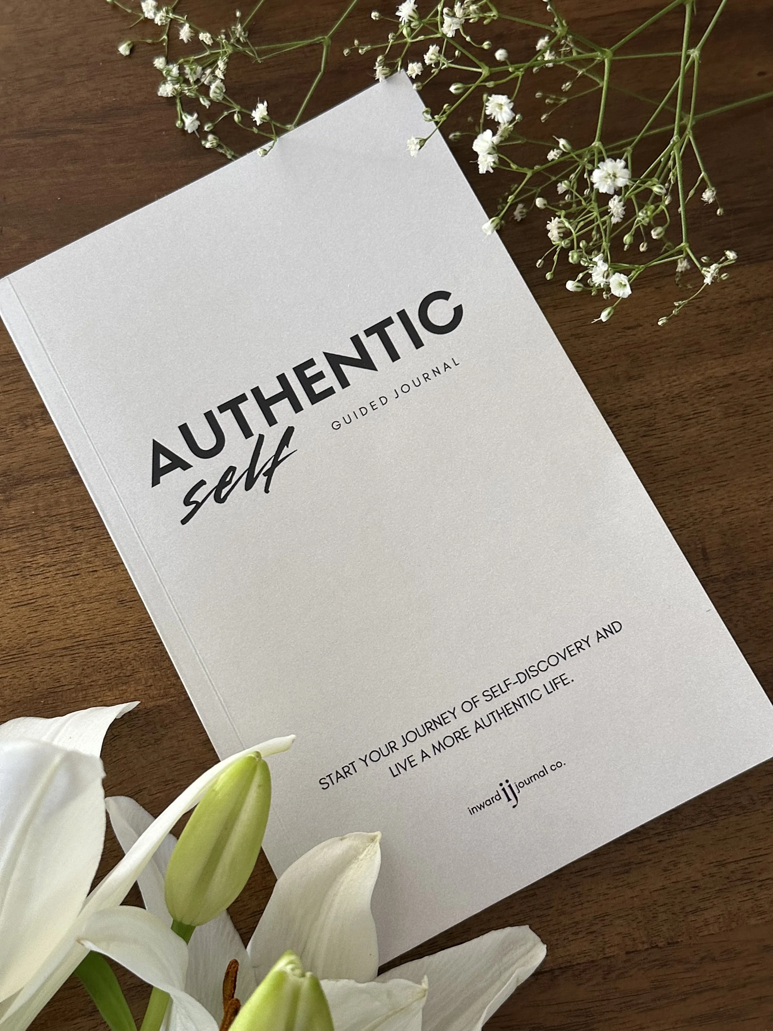 A guided journal titled 'Authentic Self' placed on a wooden surface, surrounded by white lilies and baby's breath flowers.