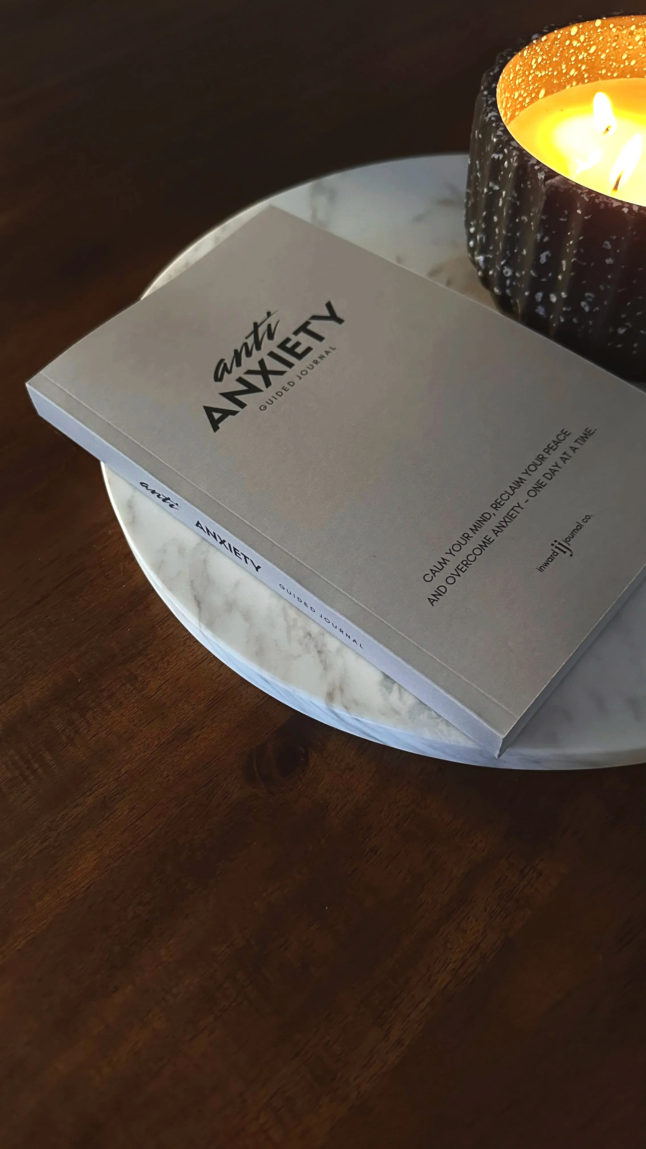 Close-up of a guided journal titled 'anti ANXIETY' resting on a marble tray beside a lit candle with black and gold exterior.