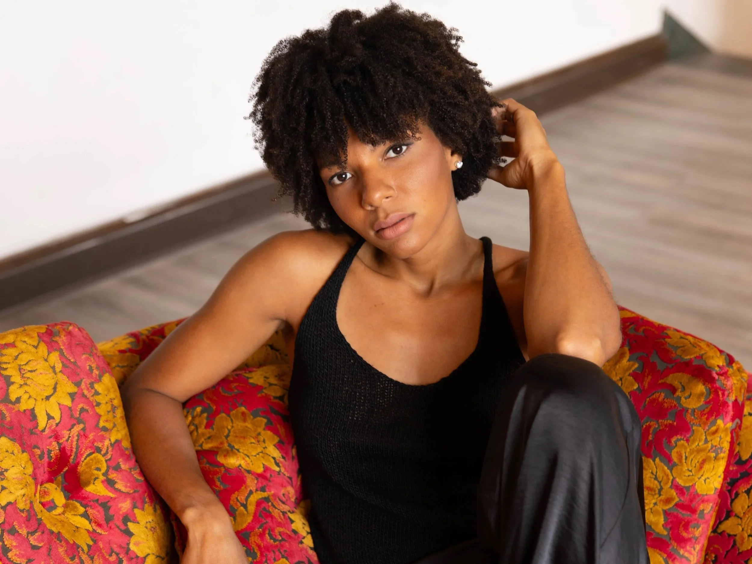 A woman with natural curly hair sitting on a colorful floral couch, wearing a black tank top and black pants, with her right arm resting on her head.