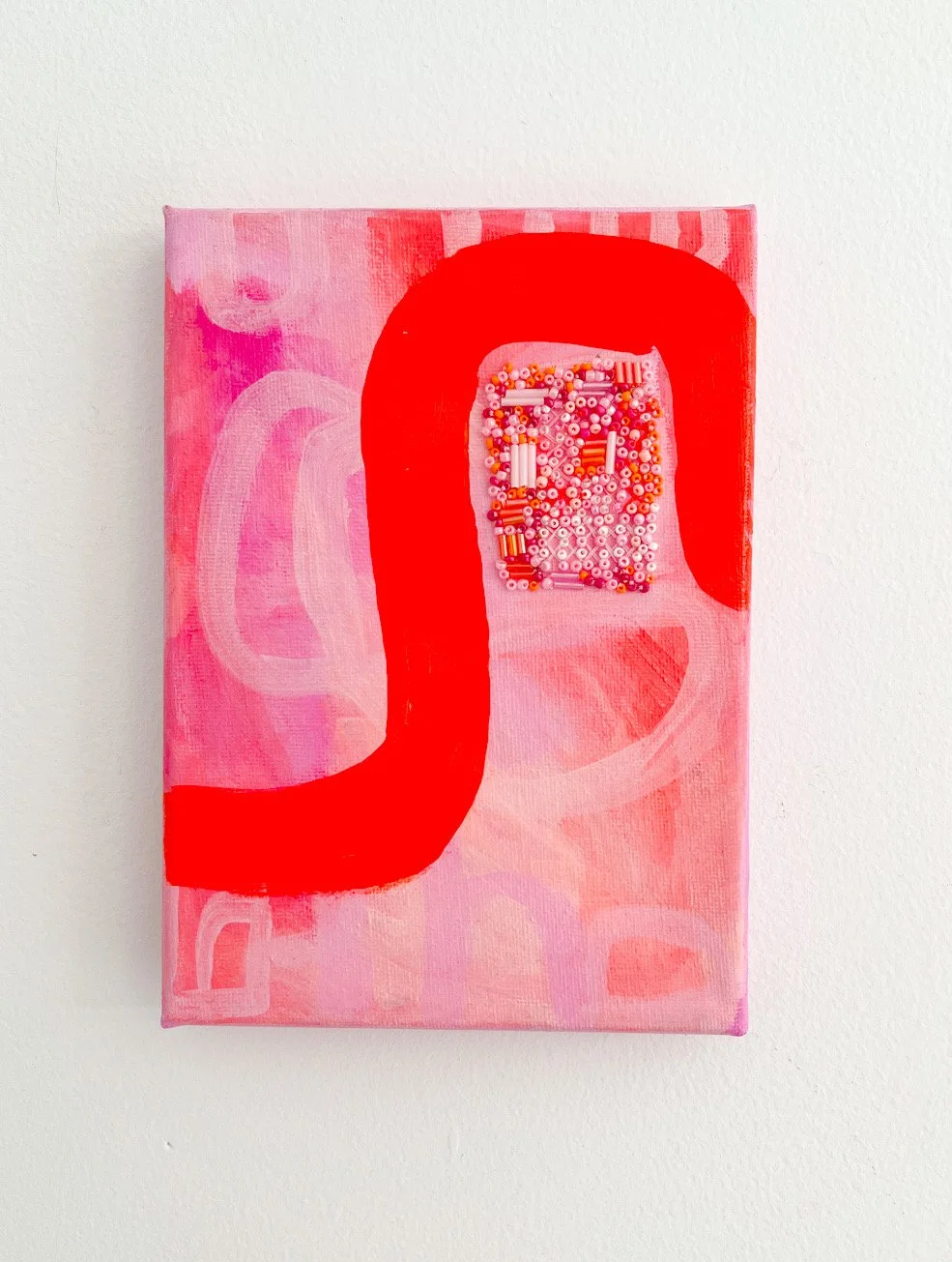 A pink abstract painting with a large red letter 'D' overlaying a background of pastel pink and white brushstrokes. The letter 'D' contains a small rectangular section filled with colorful beads.