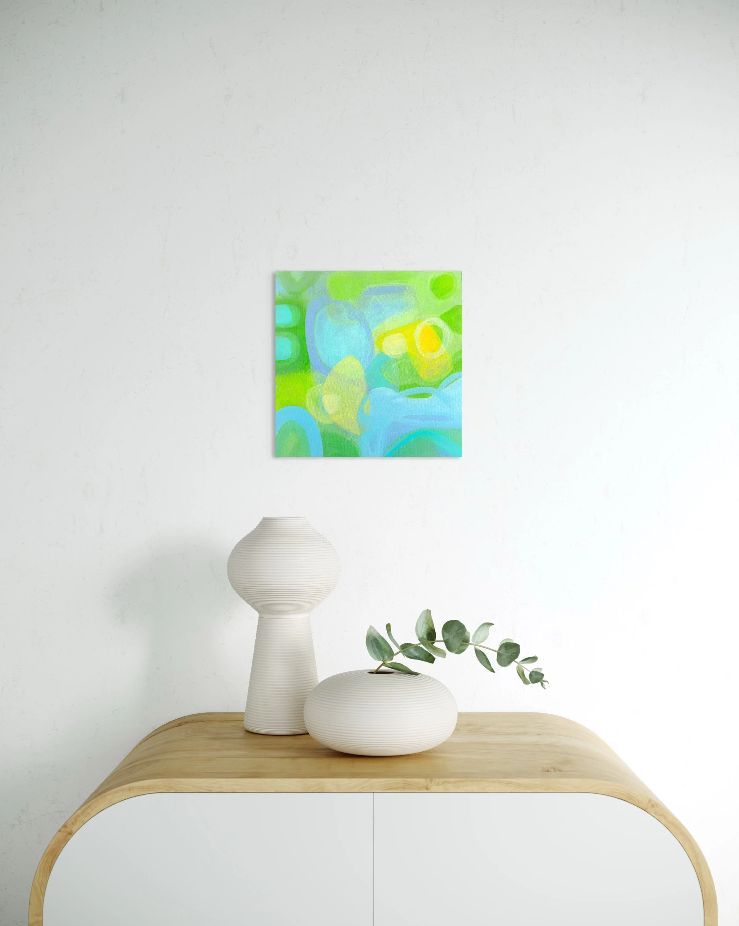 A minimalist interior with a light wooden console table holding a white vase and a sprig of eucalyptus, with an abstract colorful painting on the white wall behind.