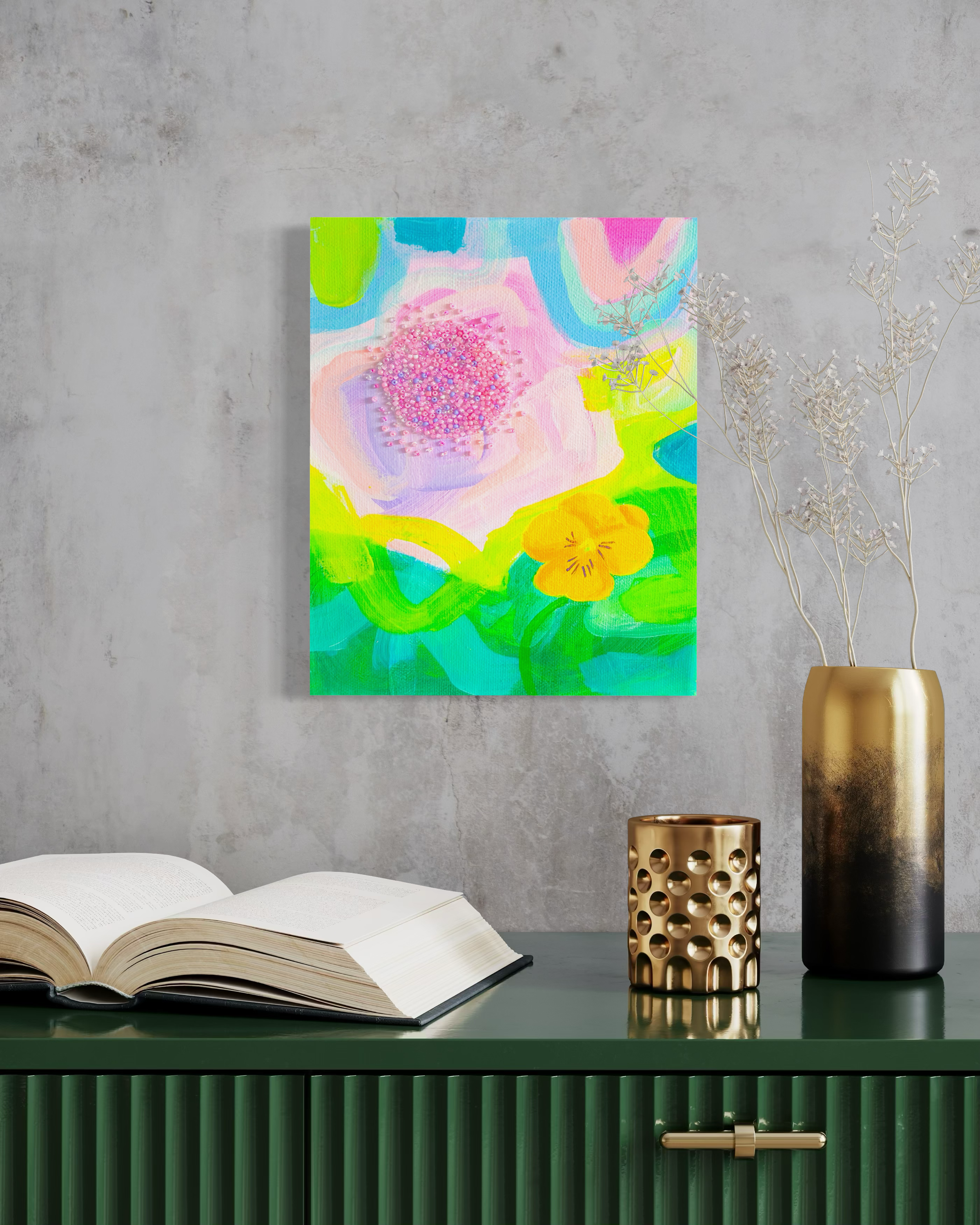 Colorful abstract flower painting on a concrete wall with a gold and black vase with dried branches, a gold candle holder, and an open book on a green textured sideboard.