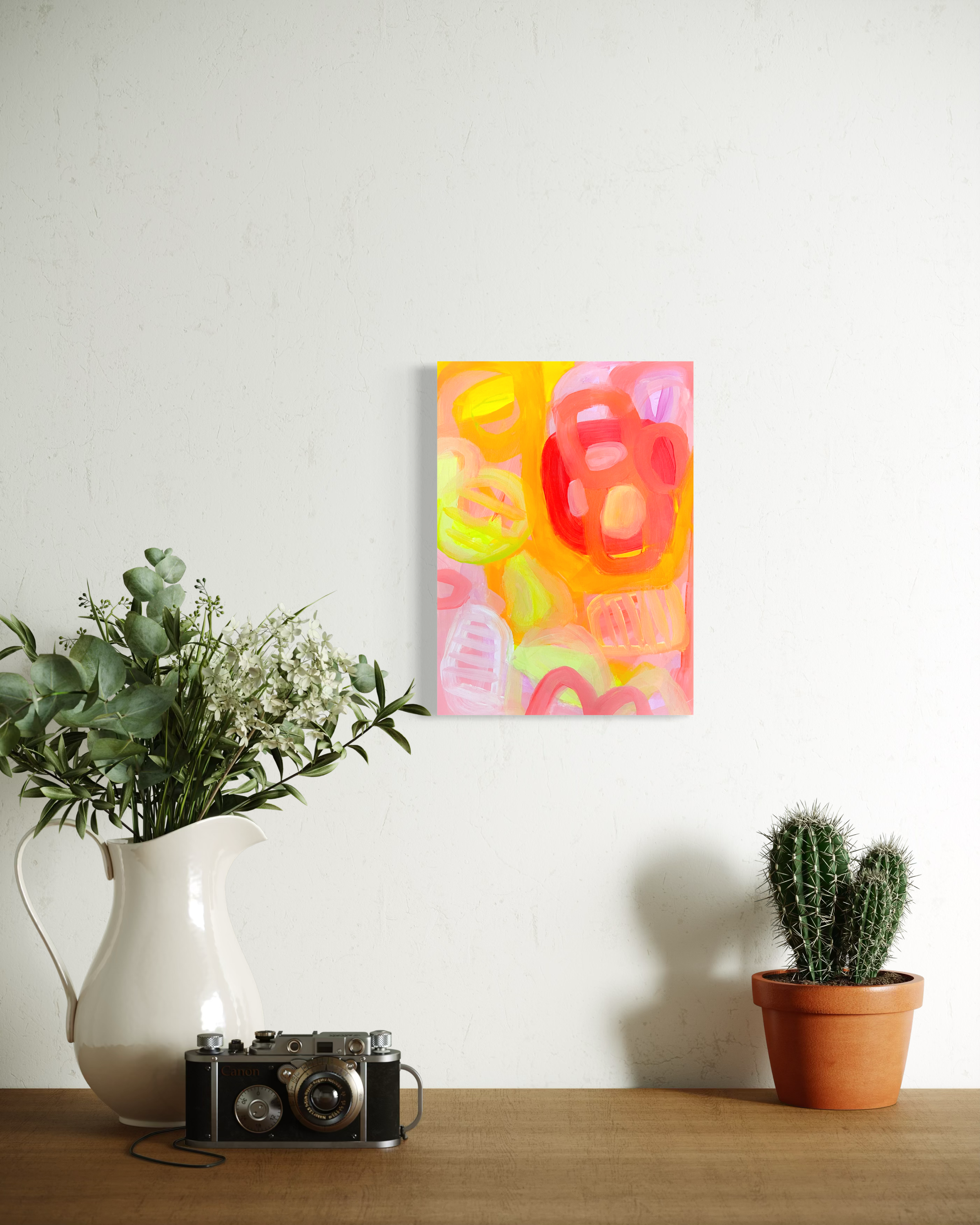 A wooden table with a white vase holding green leafy plants, a vintage black camera, a framed colorful abstract painting on a white wall, and three potted cacti.