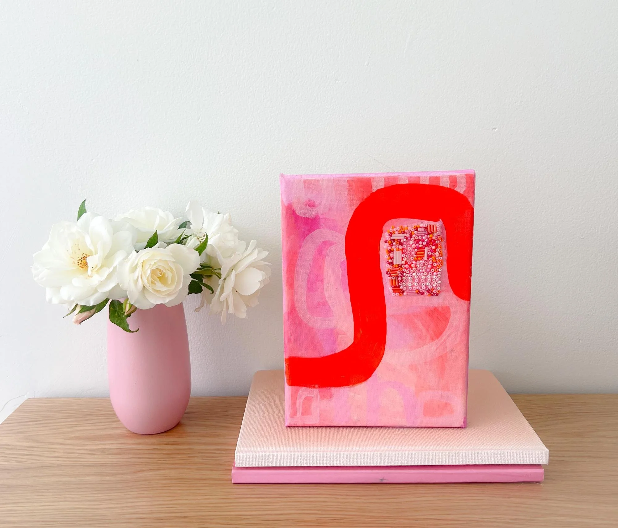 Pink vase with white flowers on a wooden surface, next to a pink book and a pink canvas with red and pink abstract art.