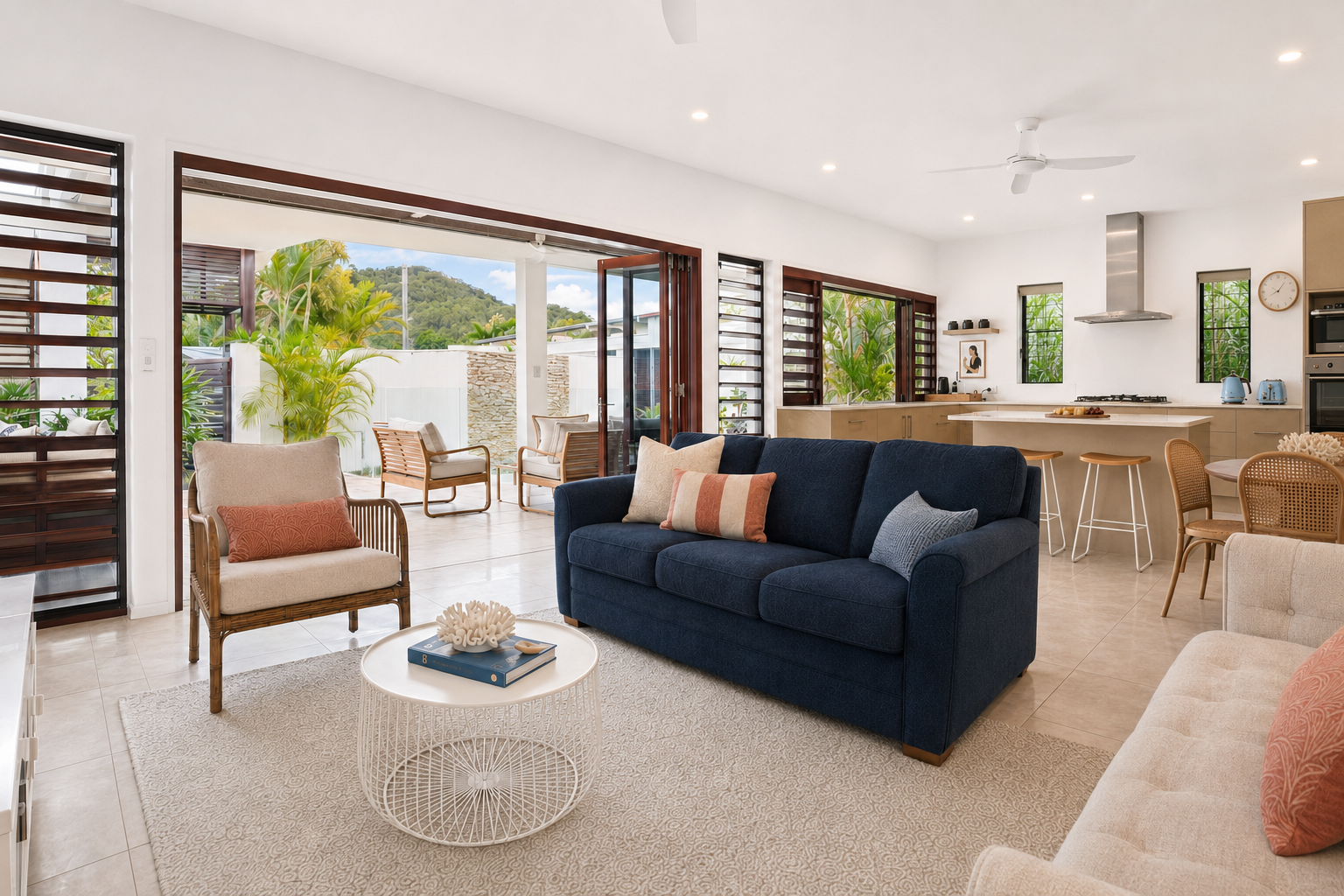 Modern living room with a navy sofa, wooden chairs, and a round coffee table. Open layout leading to a kitchen with bar stools and dining area. Large windows and a sliding door to a patio with plants.