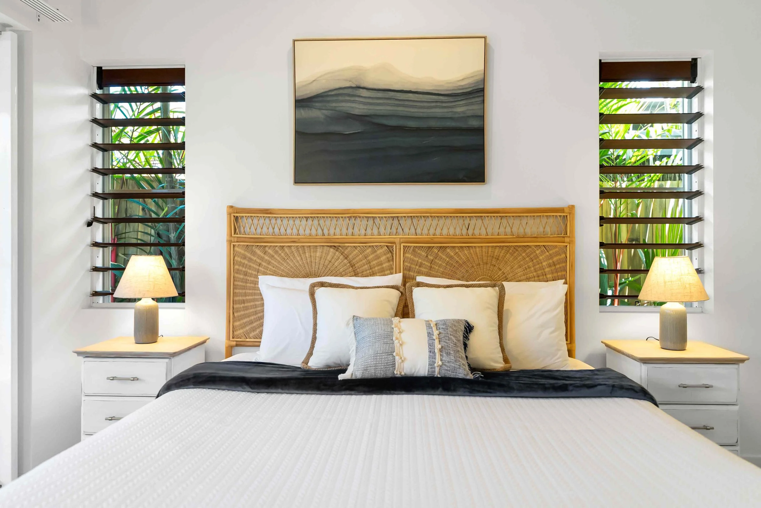 Beautiful modern bedroom with a king size rattan headboard from Mango Interiors, coastal bedside tables with lamps, abstract wall art, and window shutters.