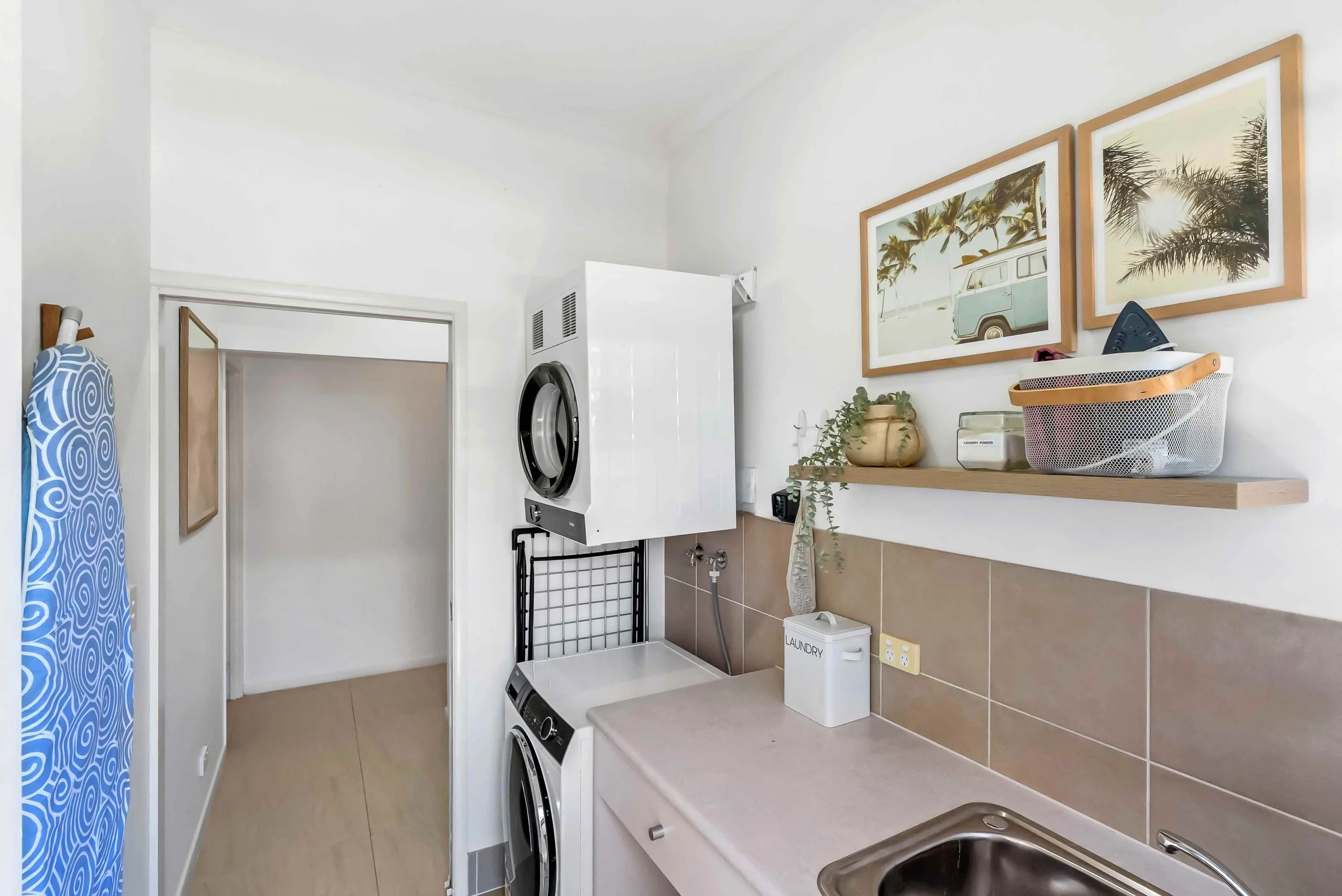 Laundry room with beige cabinets, stainless steel sink, tiled back splash, shelf with laundry tin and basket, framed pictures,coastal blue door with glass window and folding drying rack.
