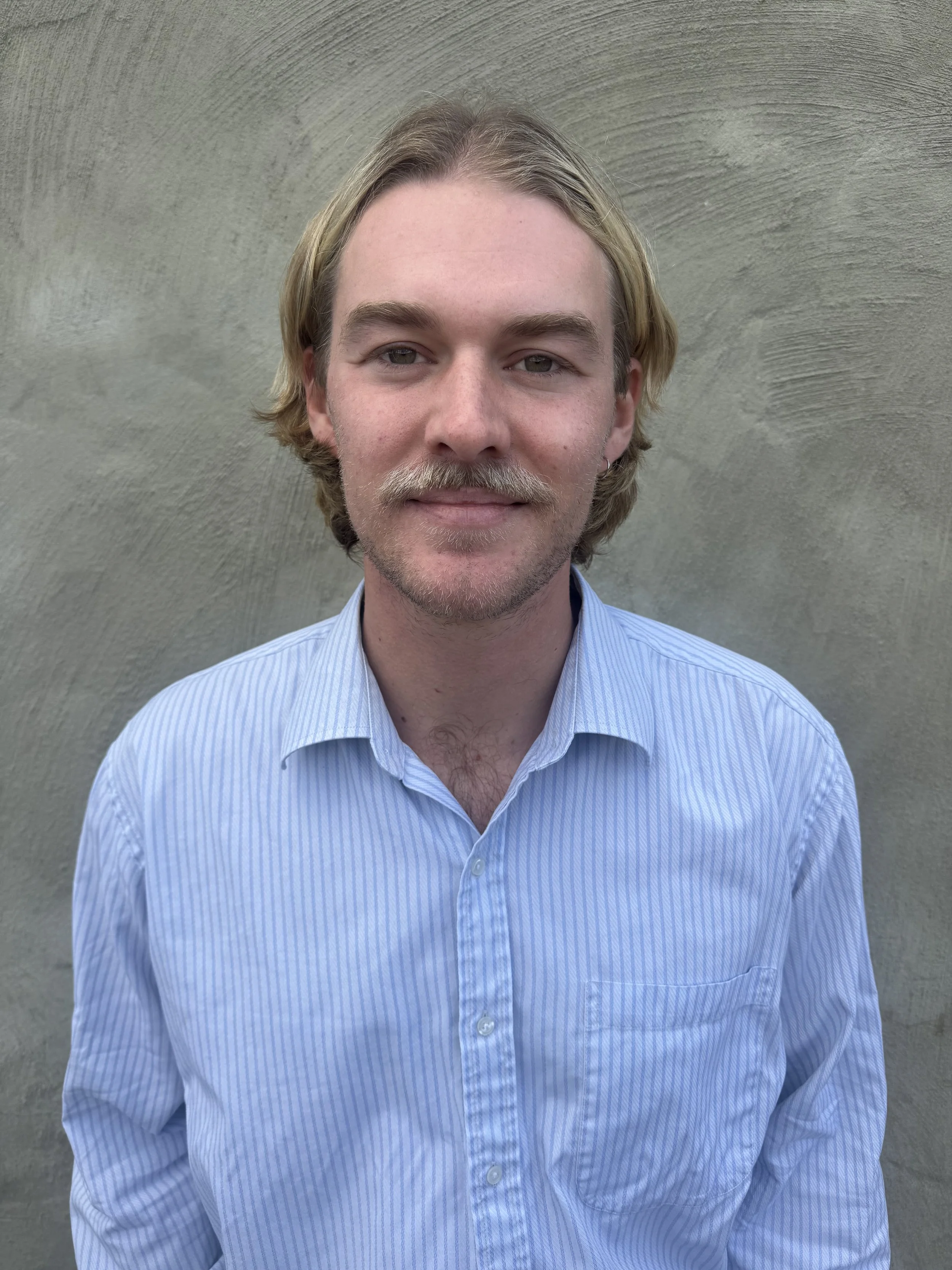 A portrait of Neuropsychology Registrar Liam Dunne. He presents as a young man with blond hair and a mustache, wearing a light blue striped button-up shirt, standing in front of a plain gray wall.