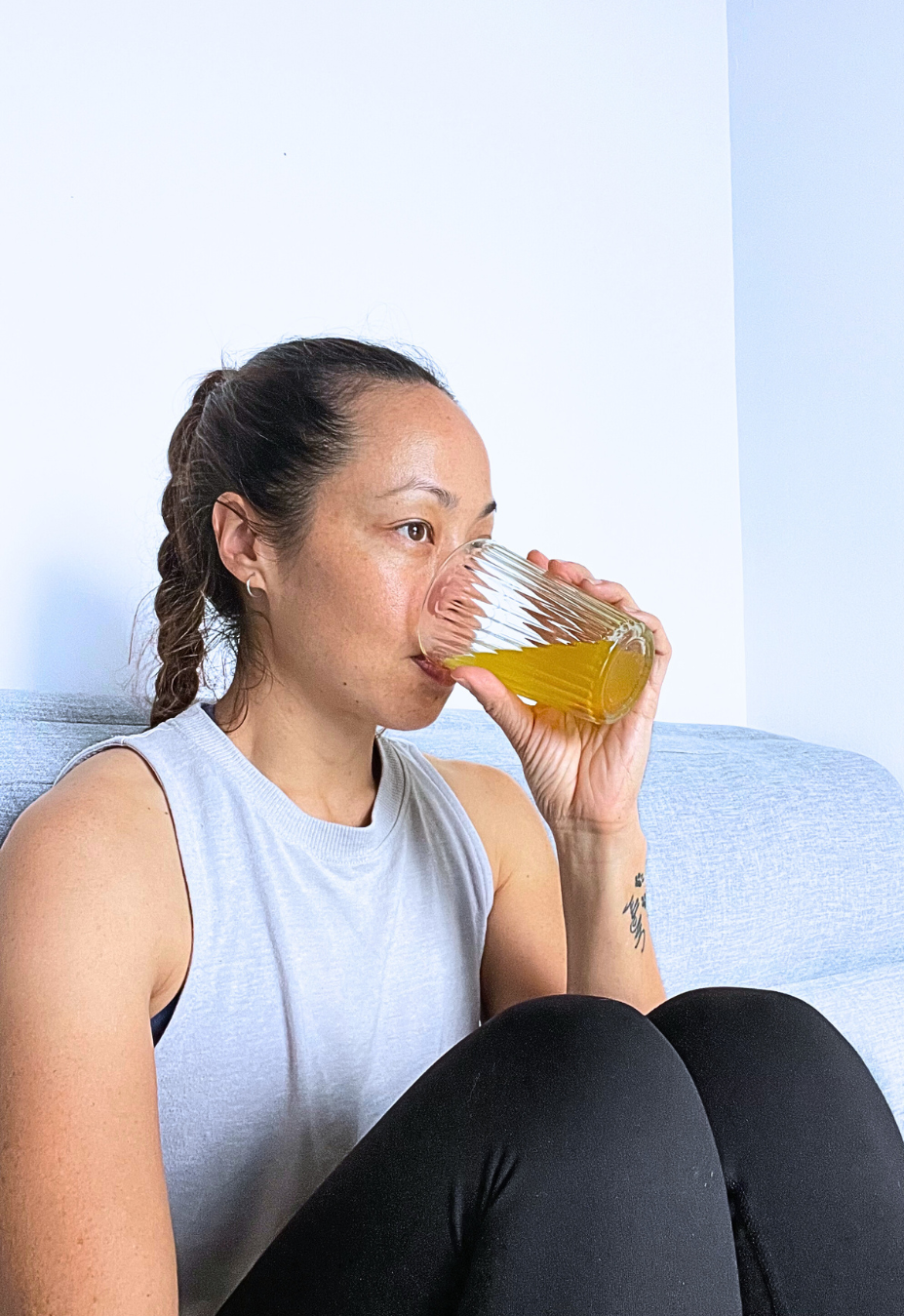 Woman with a braid wearing a light sleeveless shirt, sitting on a couch, drinking a yellow beverage from a textured glass.