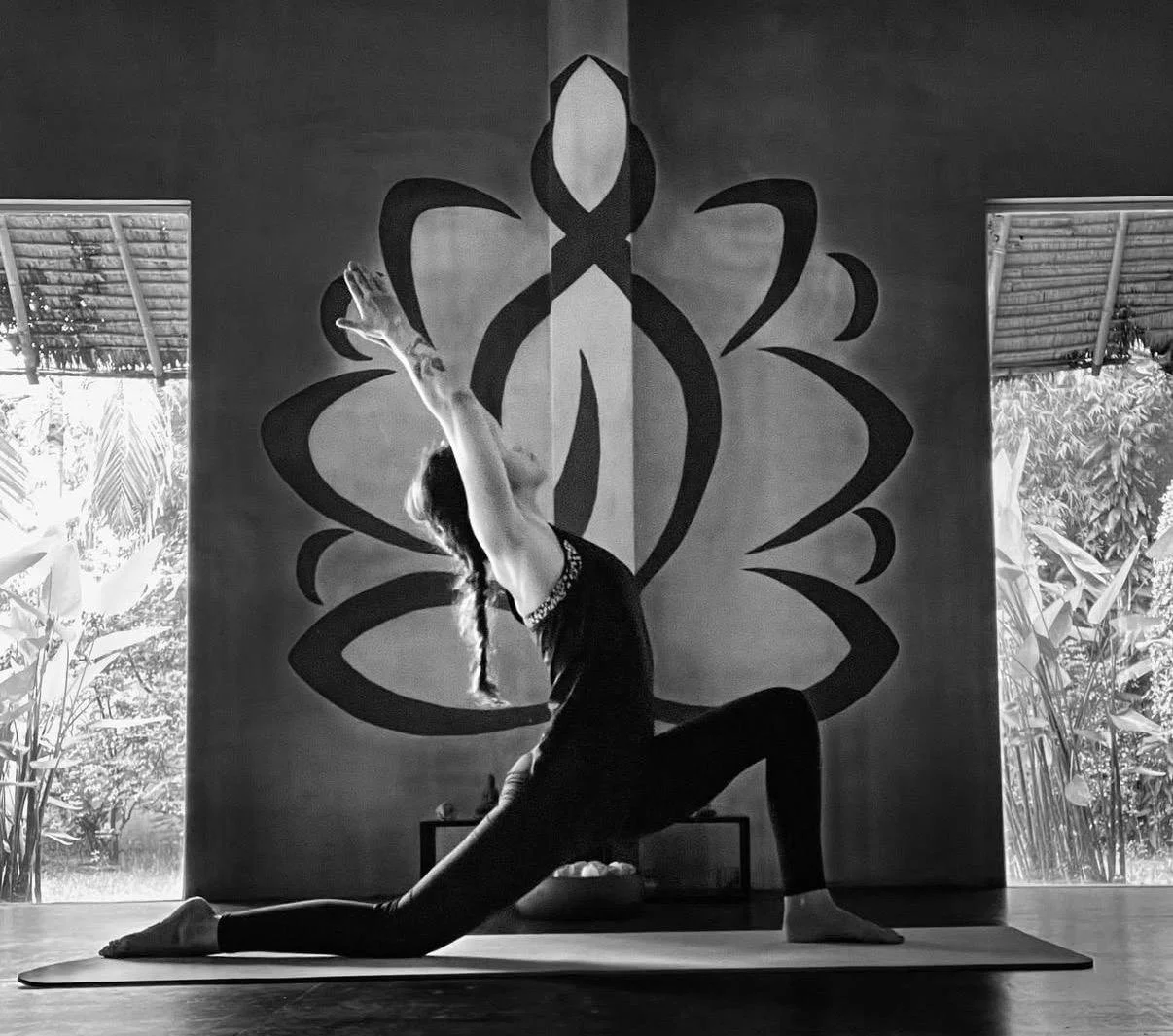 A woman practicing yoga in a lunge pose with arms raised overhead in a room with large windows showing outdoor greenery, with a decorative wall hanging behind her.