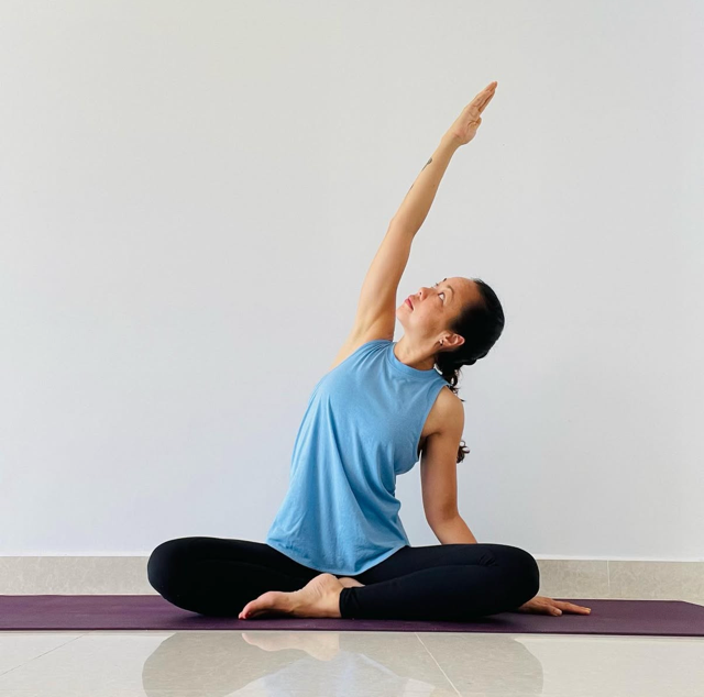 Woman in blue tank top and black leggings practicing yoga on purple mat in front of plain white wall, sitting cross-legged and reaching with her right arm up and to the side