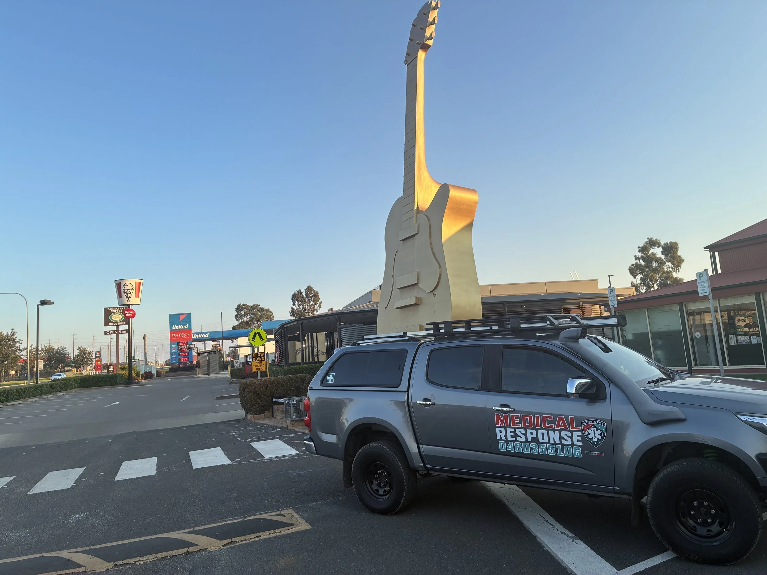 Large guitar-shaped sign on top of a vehicle parked outside a building, with fast-food and gas station signs in the background.