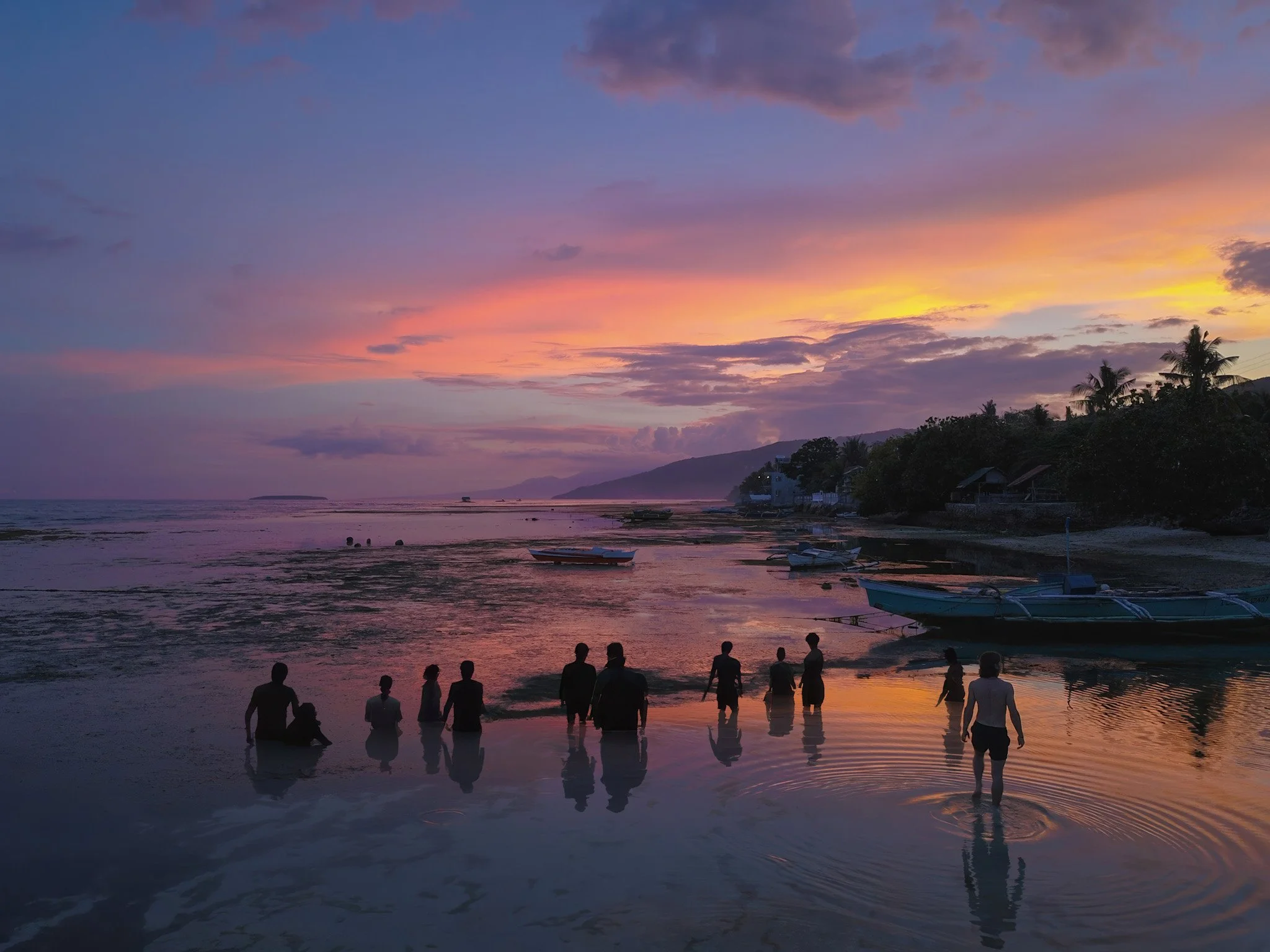 Group of people in the sea at sunset with boats on the beach, trees, and mountains in the background. Cebu, Philippines.