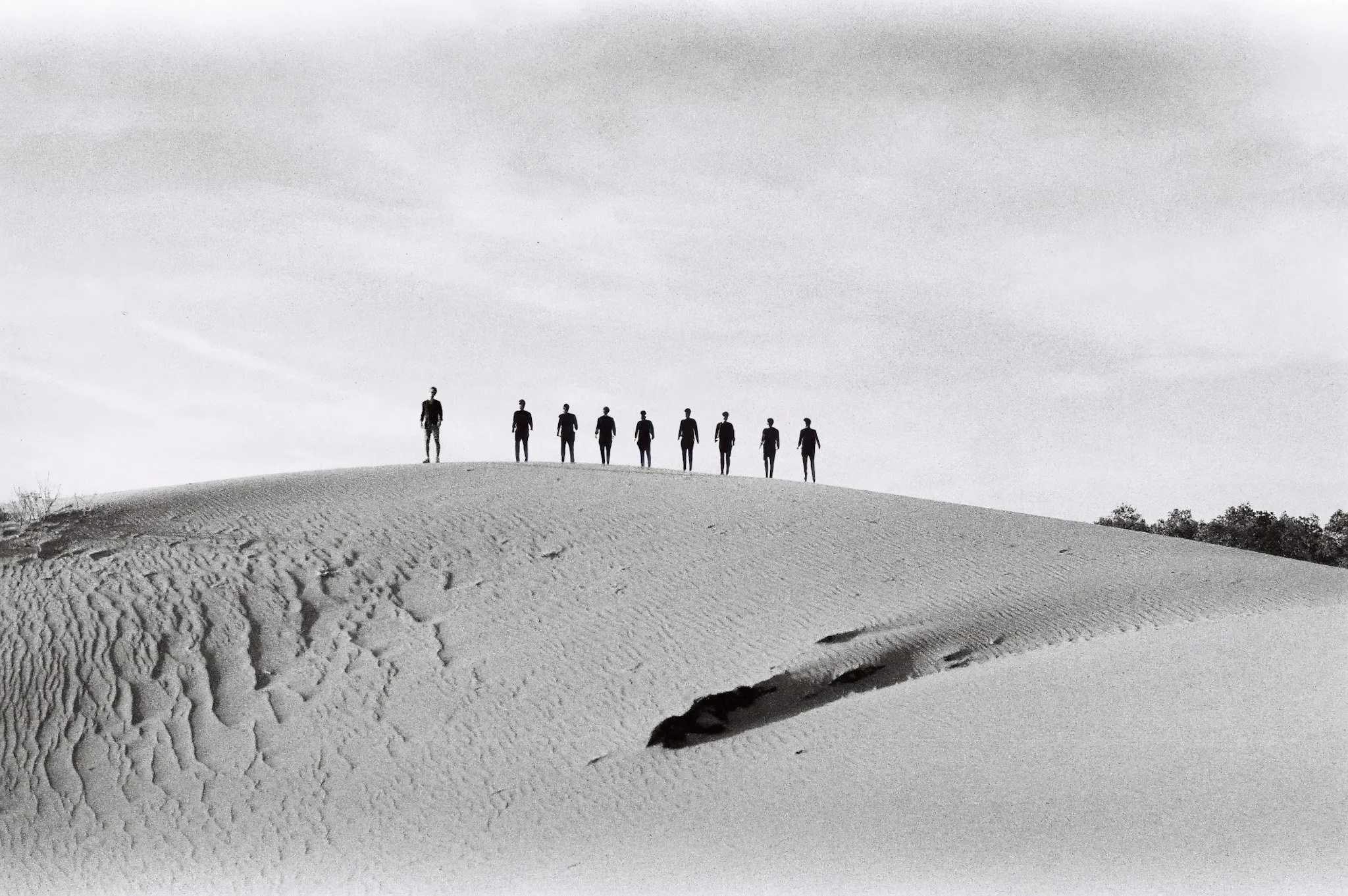 Black and white film photograph of eight silhouettes of men standing on a sand dune, film grain, viewed from below, with a cloudy sky in the background. Mui Ne, Vietnam.
