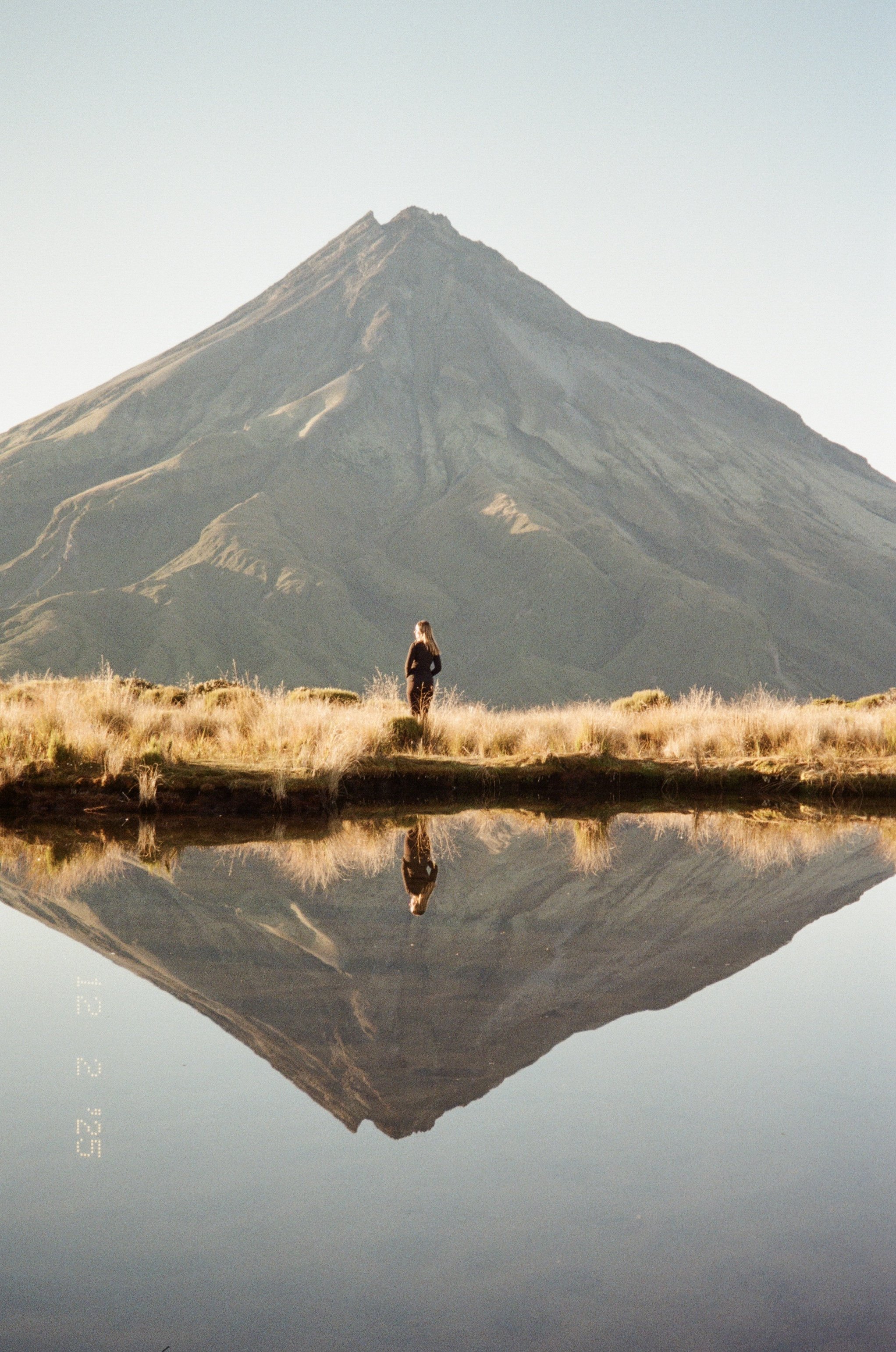 Person standing near a reflective lake with a large mountain in the background at sunrise.