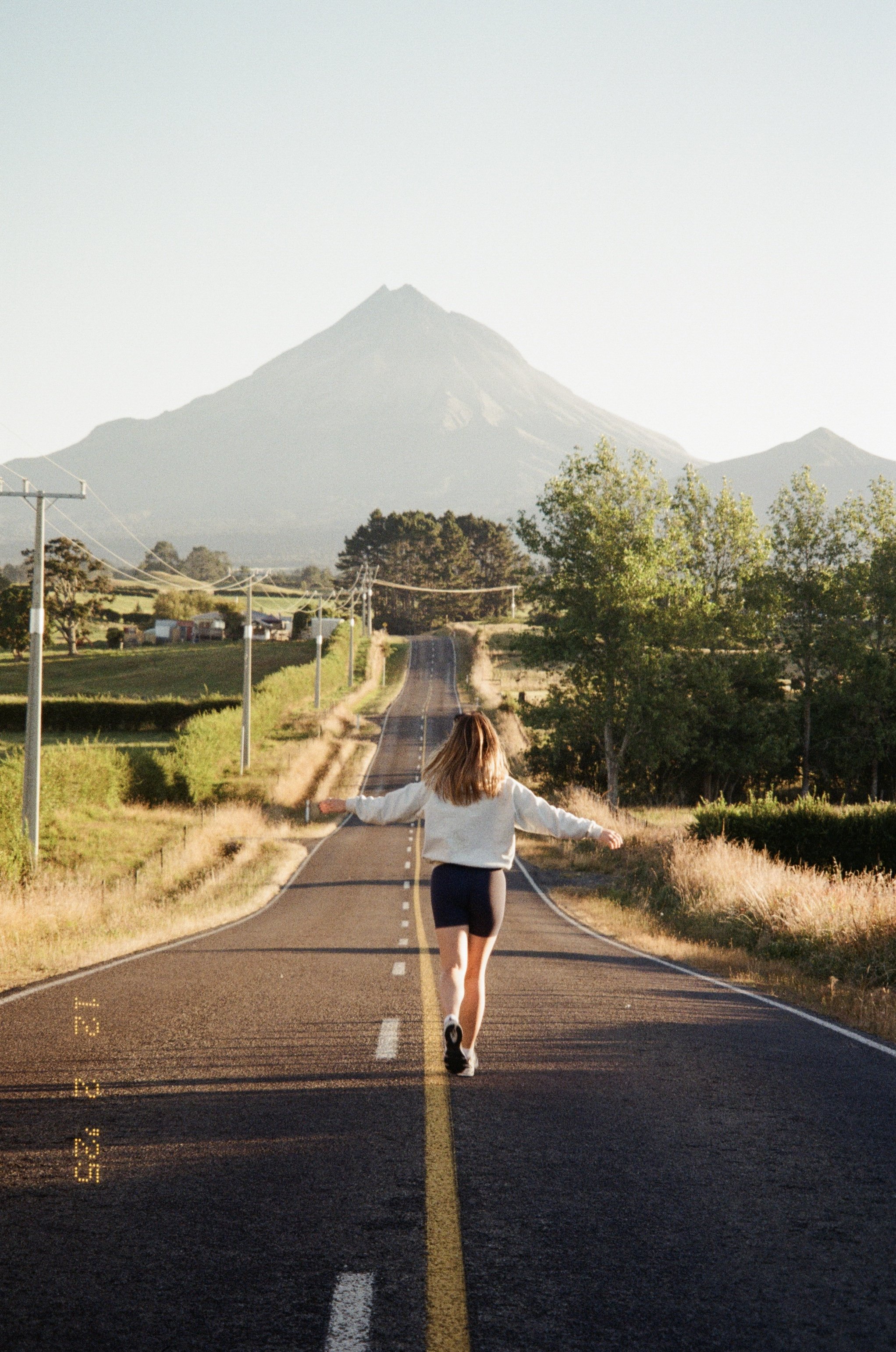 Person walking down a rural road towards a mountain, surrounded by fields and trees, on a sunny day.