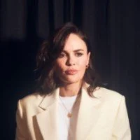 Person with brown hair wearing a white blazer against a dark background.