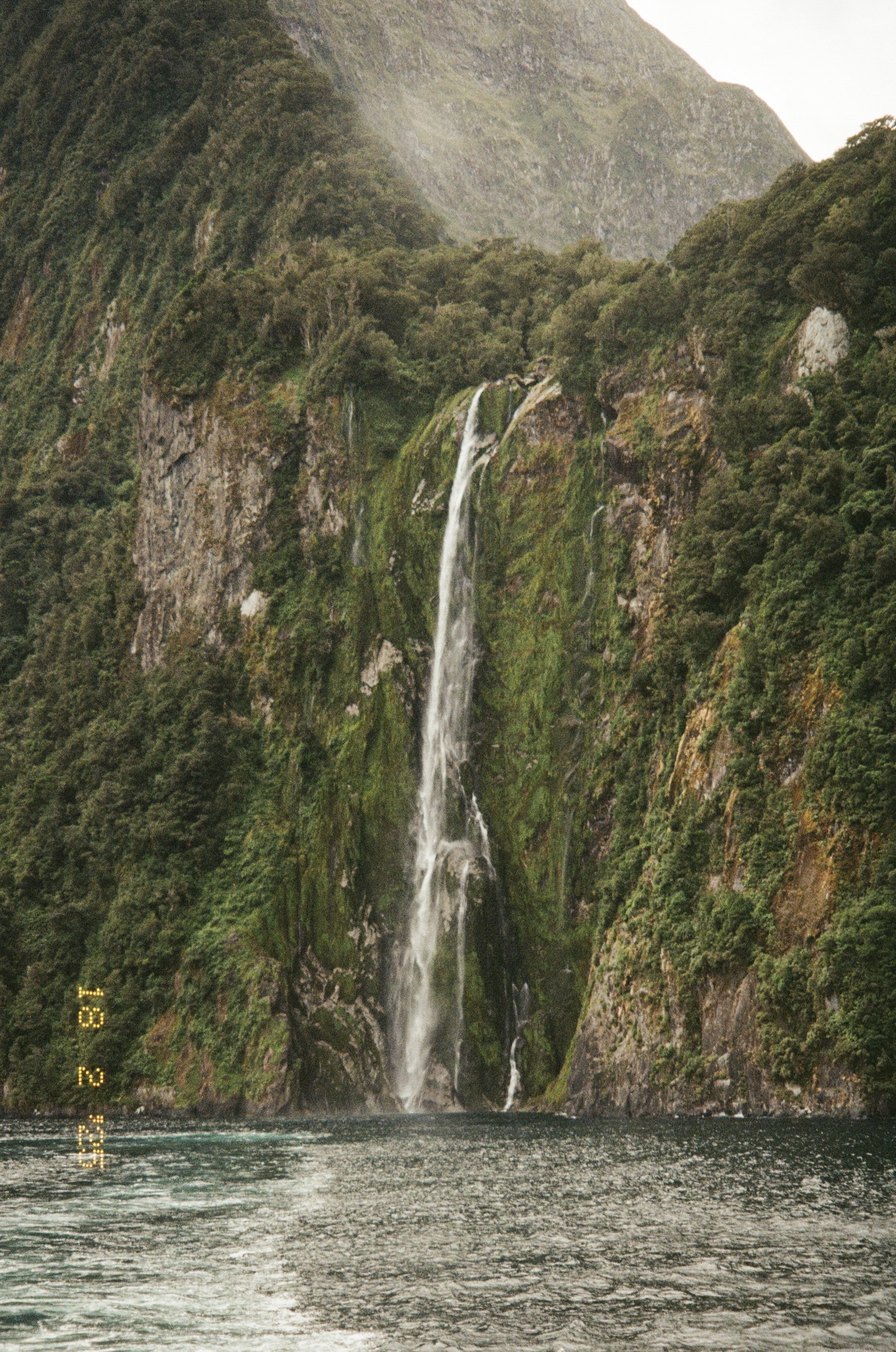 Waterfall cascading down a lush, green cliff into a body of water, surrounded by dense forest and rugged mountains.
