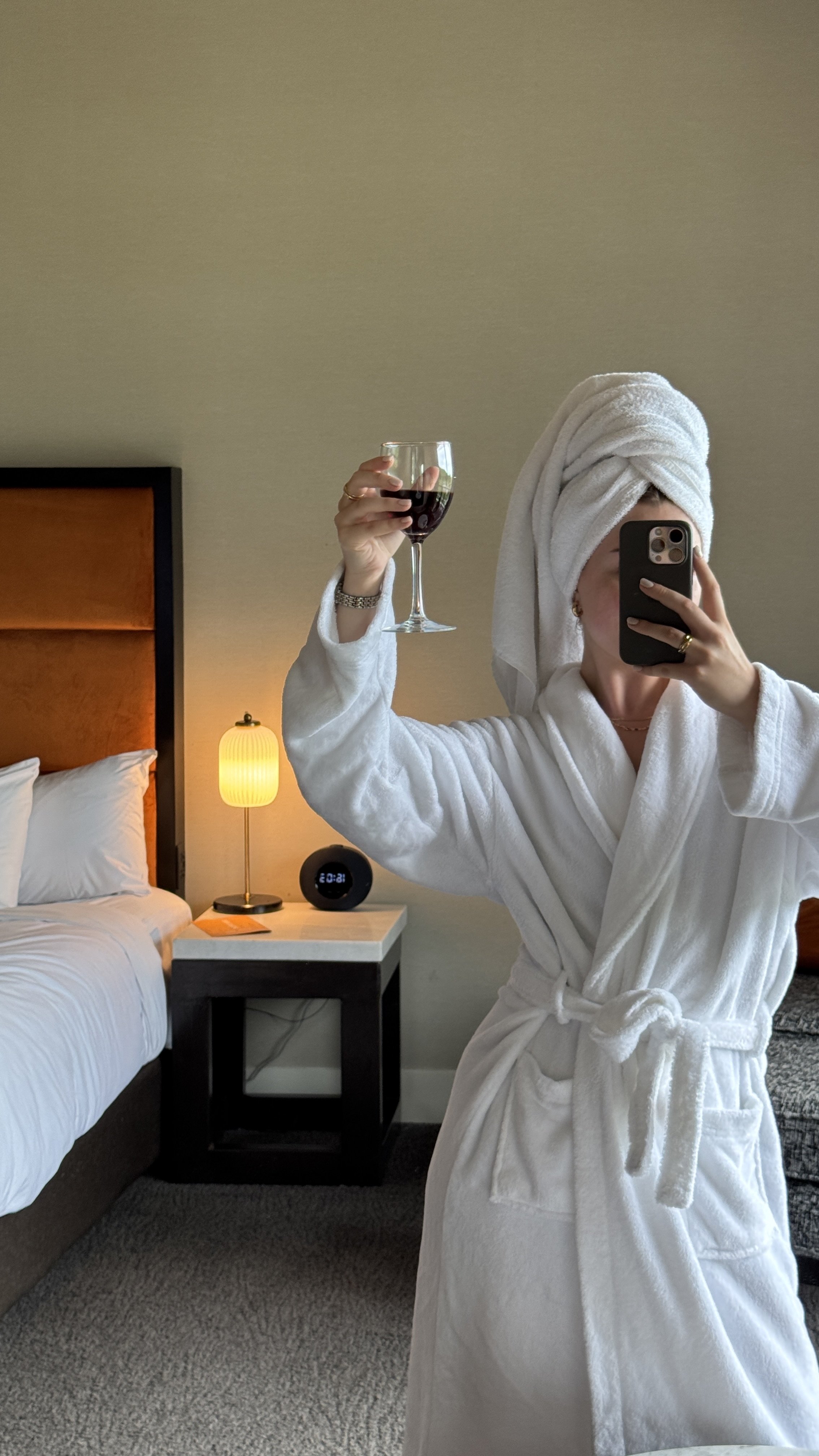 Person in robe with towel on head taking selfie, holding wine glass in hotel room.