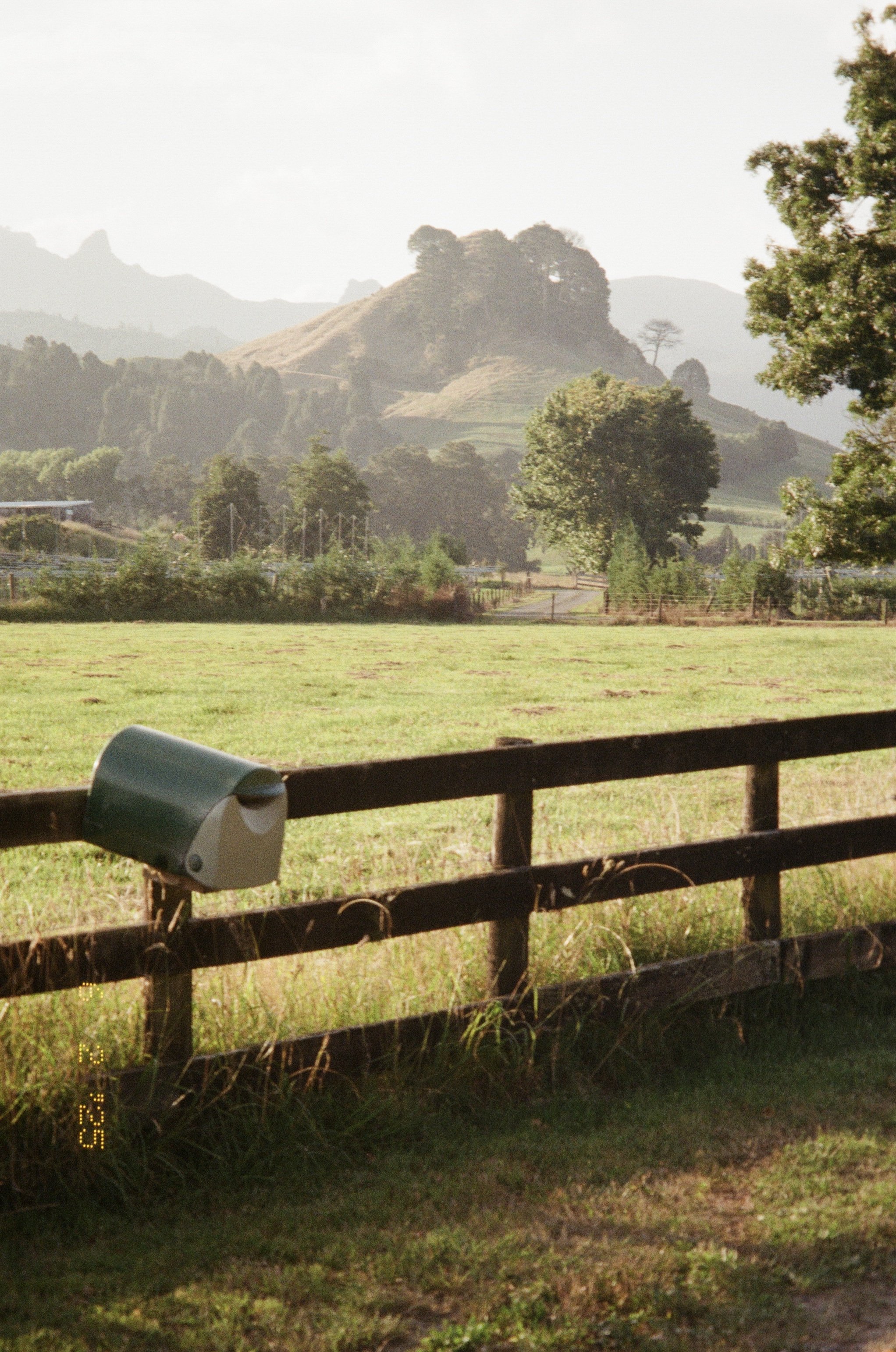 Rustic rural scene with a wooden fence, green rolling hills, trees, and distant mountains.