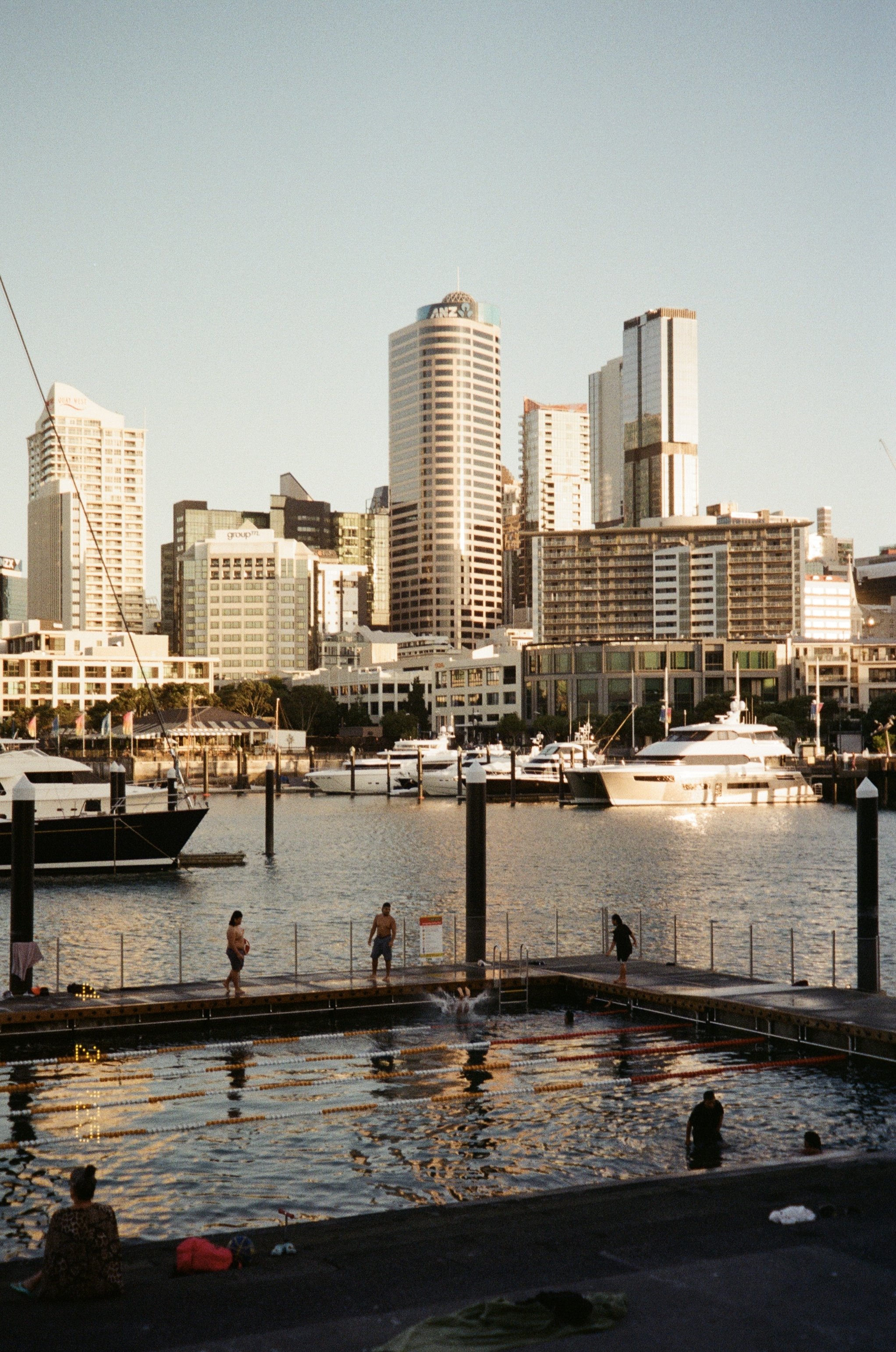 Urban waterfront scene with yachts and skyscrapers in the background, and people enjoying an outdoor pool in the foreground.