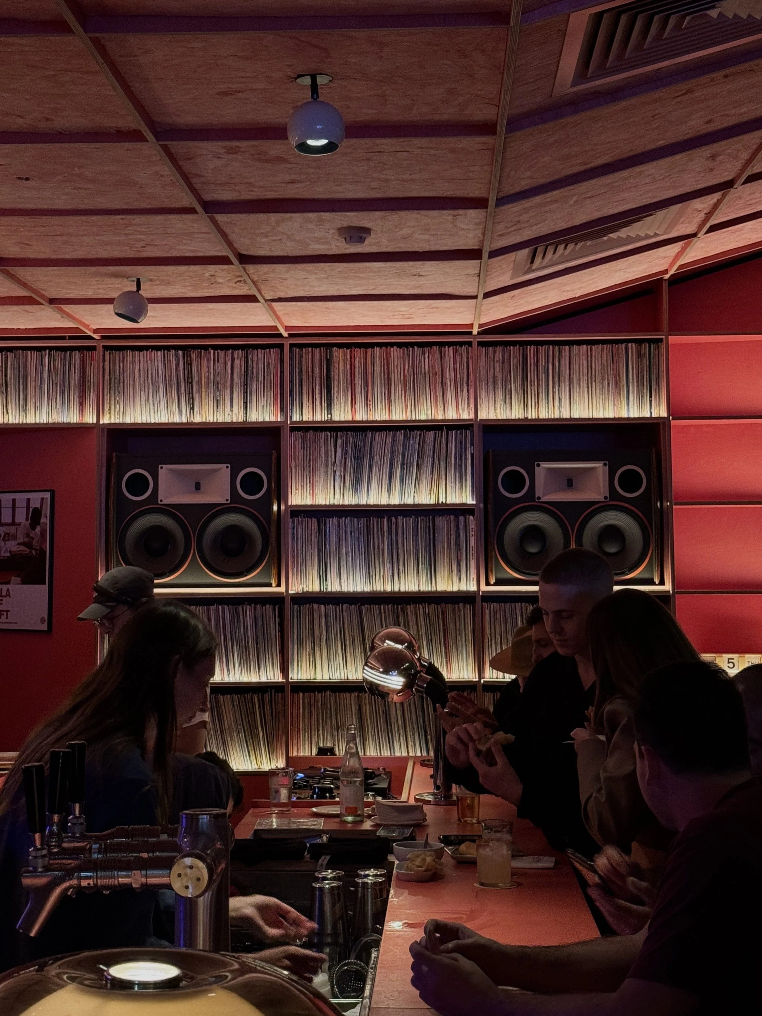 A dimly lit bar interior with patrons at the counter, featuring shelves of vinyl records and large speakers on the wall, creating a cozy and retro atmosphere.