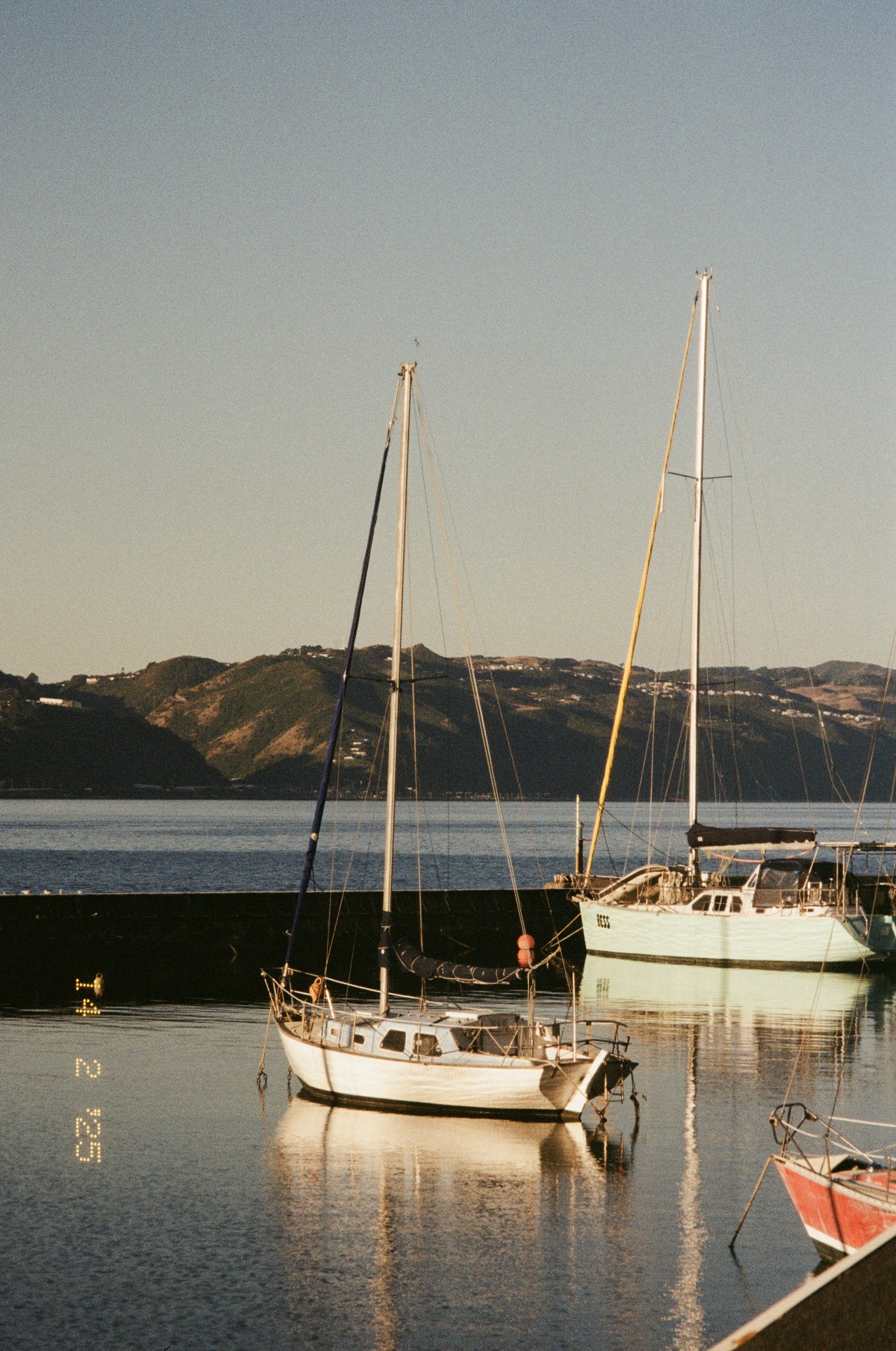 Sailboats anchored in a calm harbor with distant hills and clear sky at sunset.