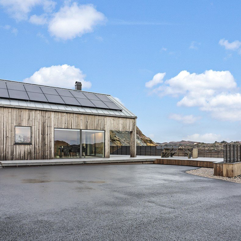 Neo hytter som enebolig. Et moderne hus med solcellepaneler på taket og store vindusflater. Stor terasse og asfaltert oppstillingsplass for biler. Huset ligger i et naturskjønt område med fjell i bakgrunnen, blå himmel og enkelte skyer.