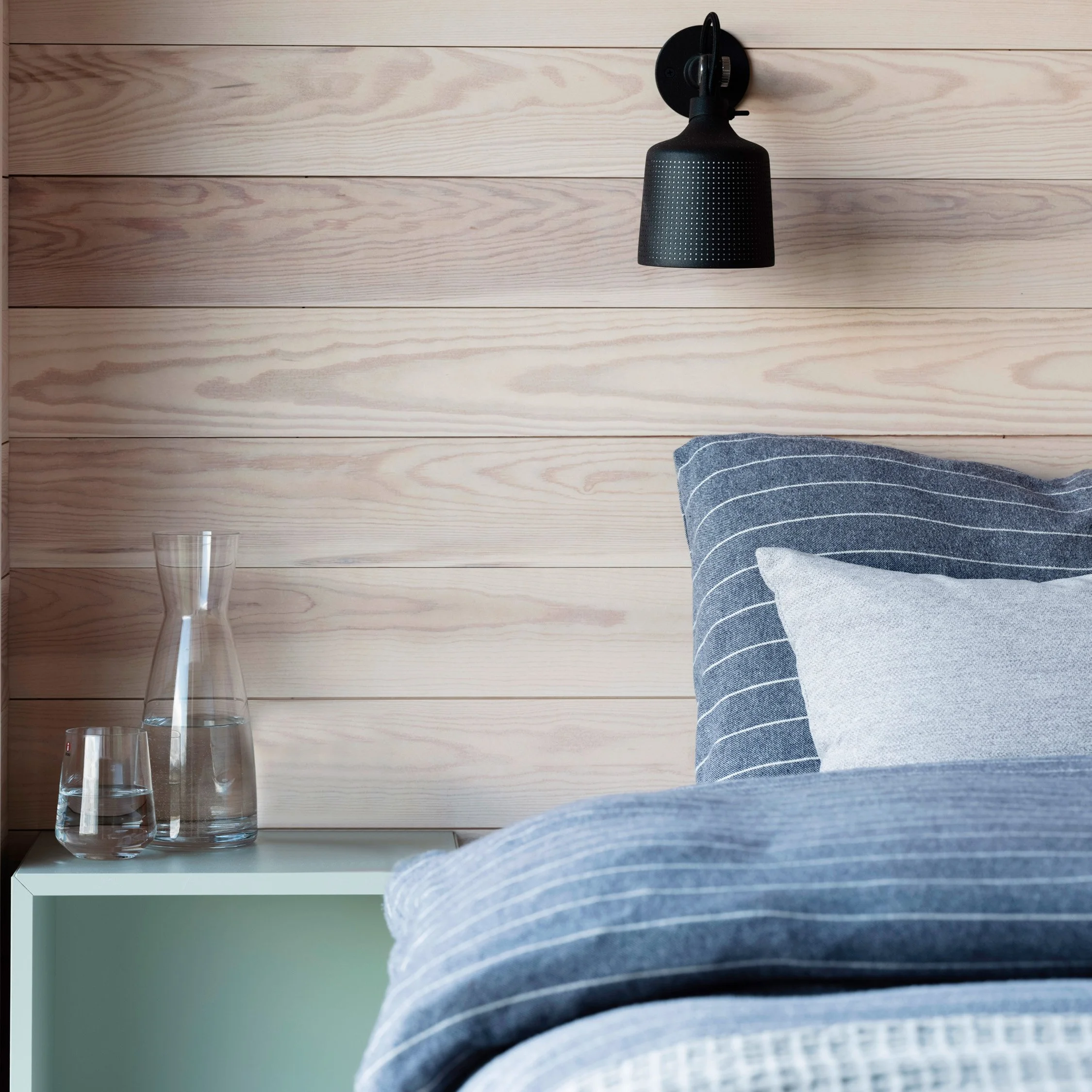 Soverom med liggende trepanel, oppredd seng med blått sengetøy og en lys pute, grønt nattbord og en sort vegglampe over senga.