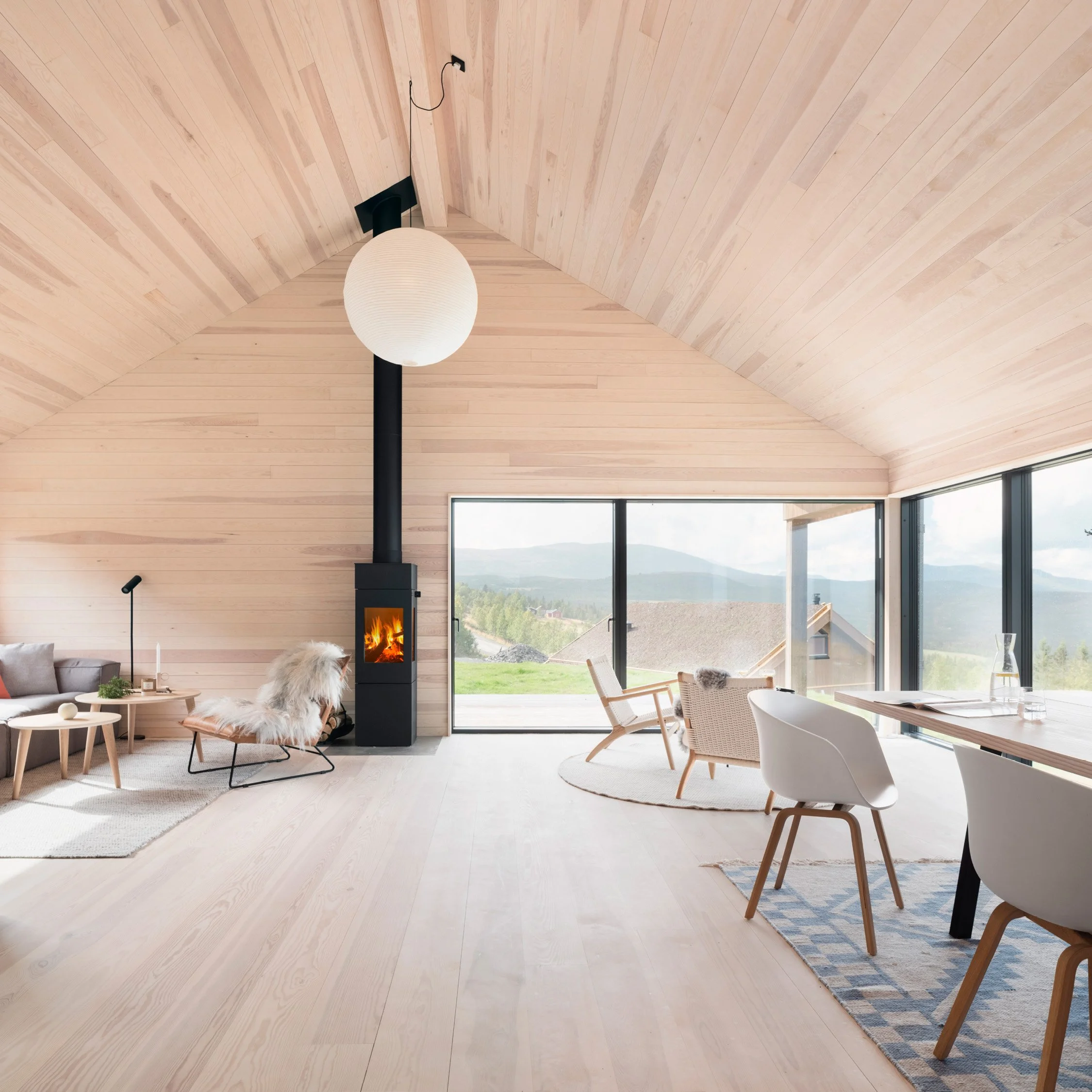 Stue med lyst panel på vegger og tak, peis, store vinduer med utsikt over landskap, moderne belysning og komfortable stoler.
