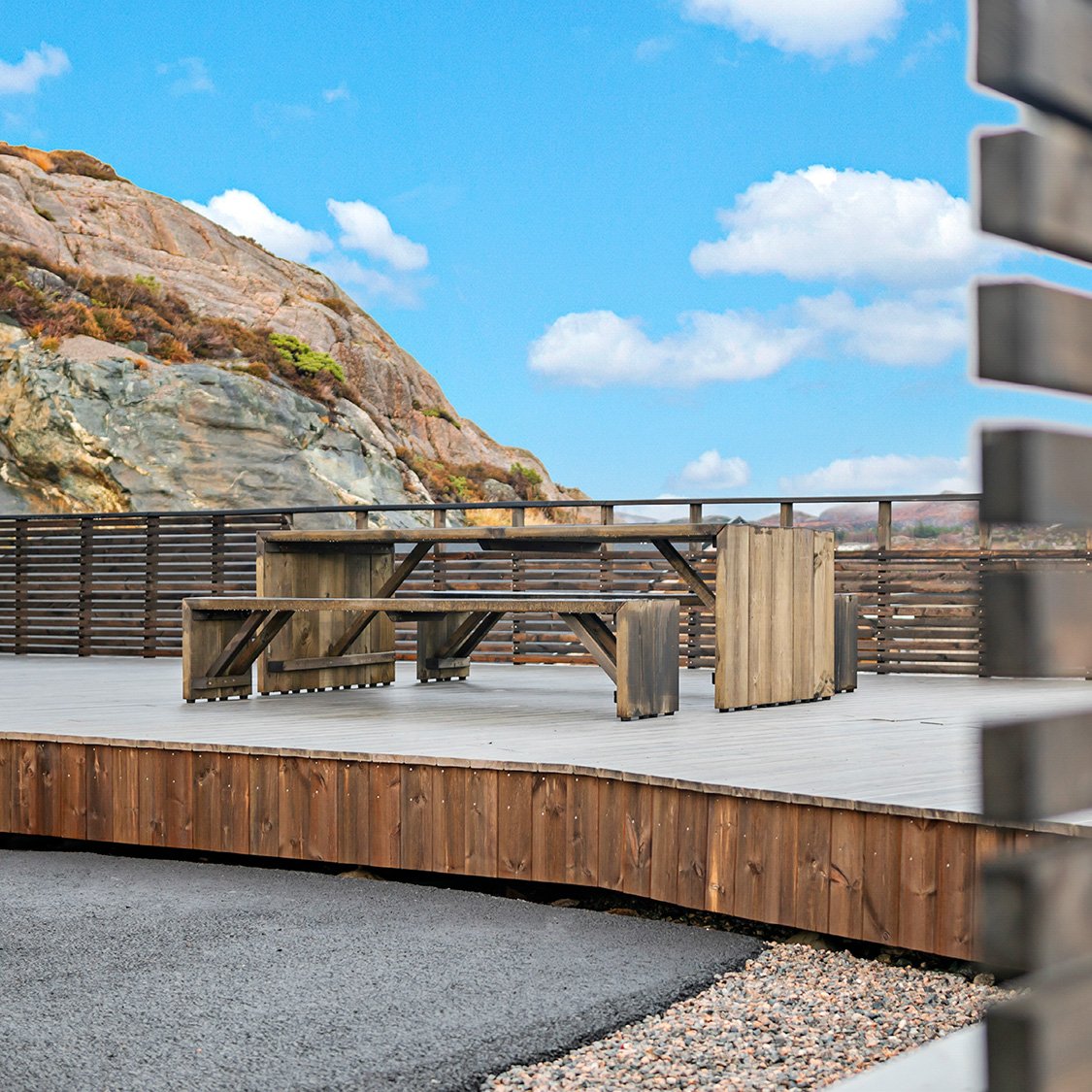 Neo hytter som enebolig. Utendørs bilde av terasse, med utebord og benk i samme materiale som kledning på vegg og terrasse. I bakgrunnen ser man fjell og blå himmel med enkelte skyer.