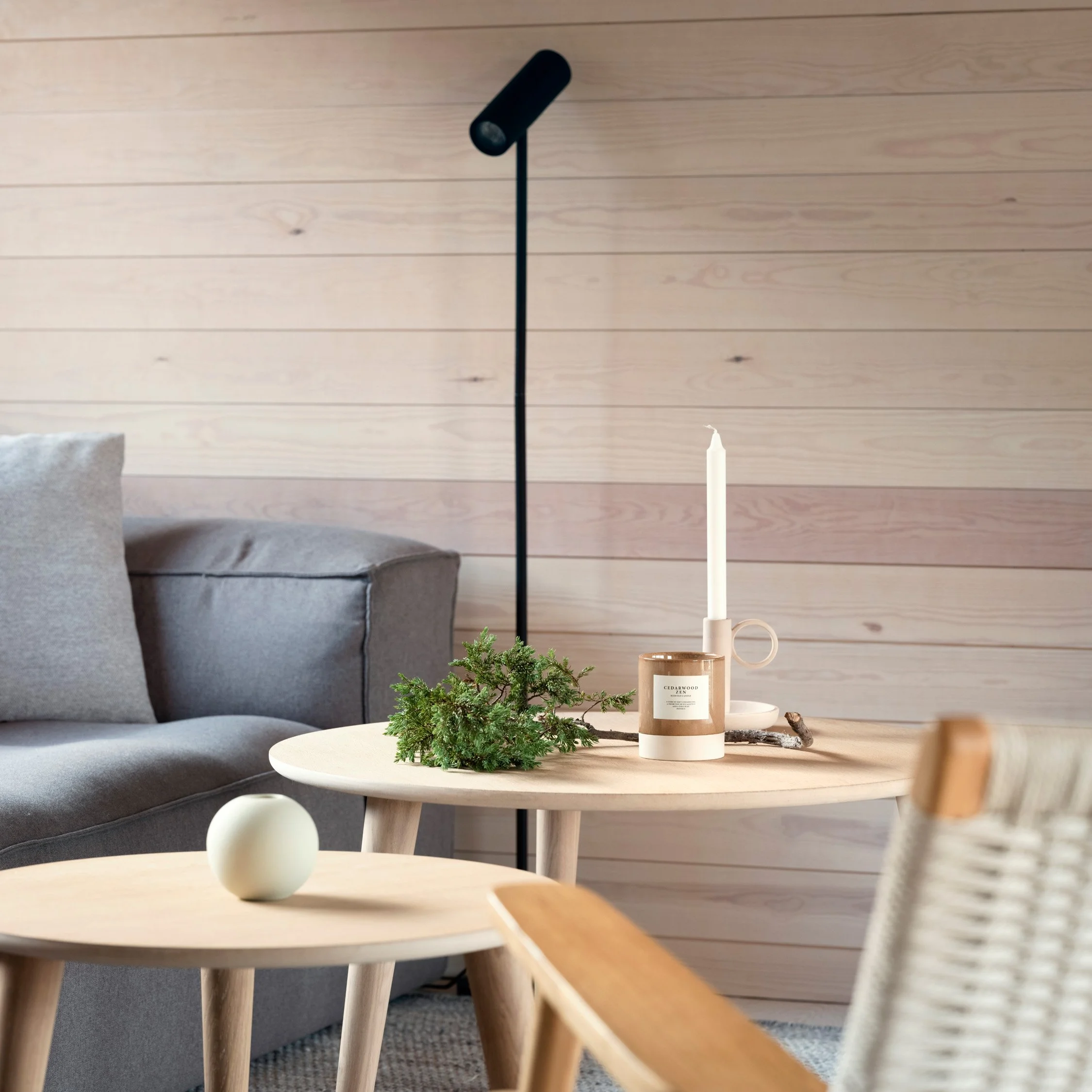 Stue med grå sofa, to små bord med pynt, lysestaker og einekvist og en moderne gulvlampe. Vegg i lyst trepanel.