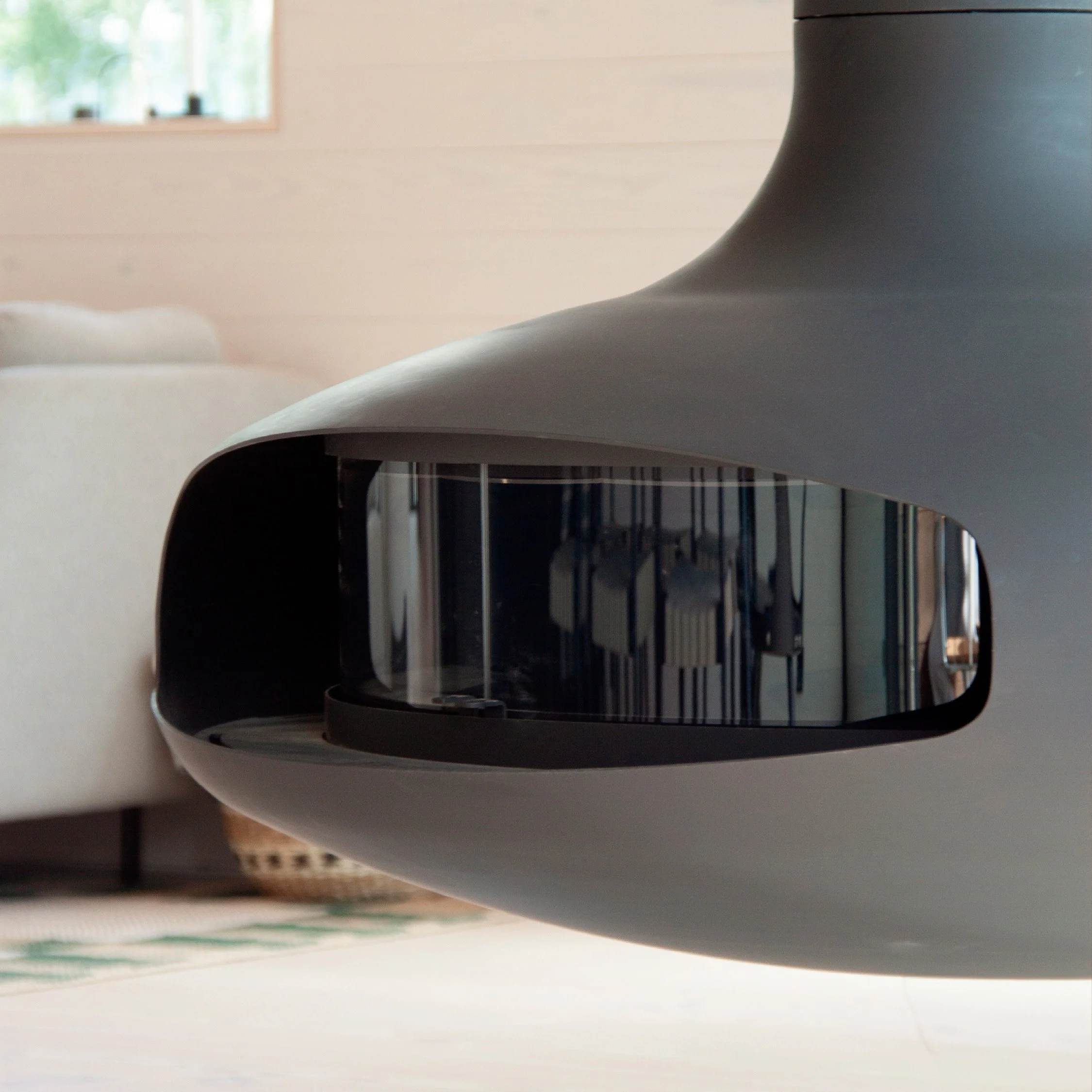 En moderne sort peisovn som henger fra taket, mot store vinduer med utsikt over grønne trær og vann i bakgrunnen.