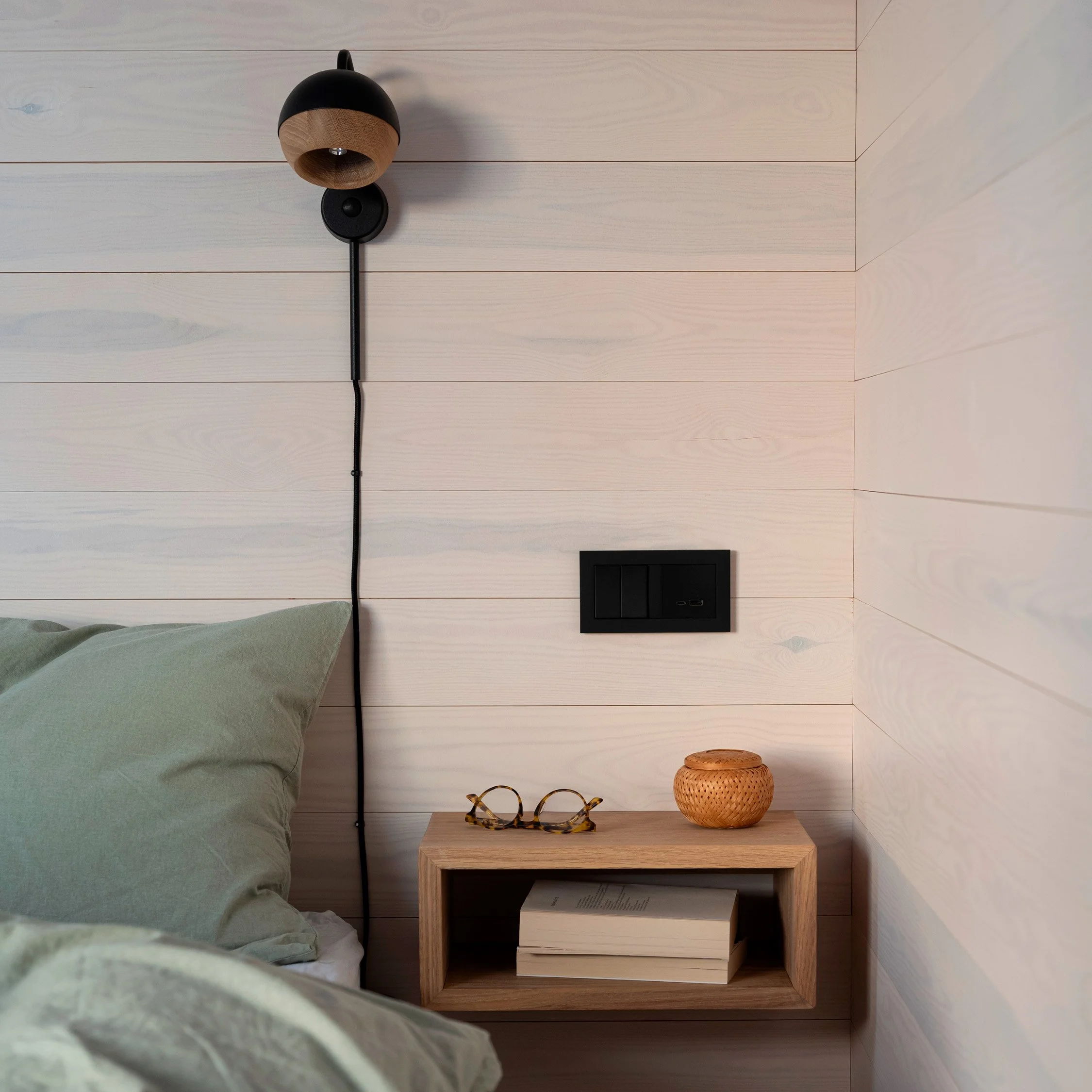 Soverom med lyst trepanel. På bildet ser man en oppredd seng med grønt sengetøy, en sort vegglampe, sort stikkontakt og et nattbord med bøker