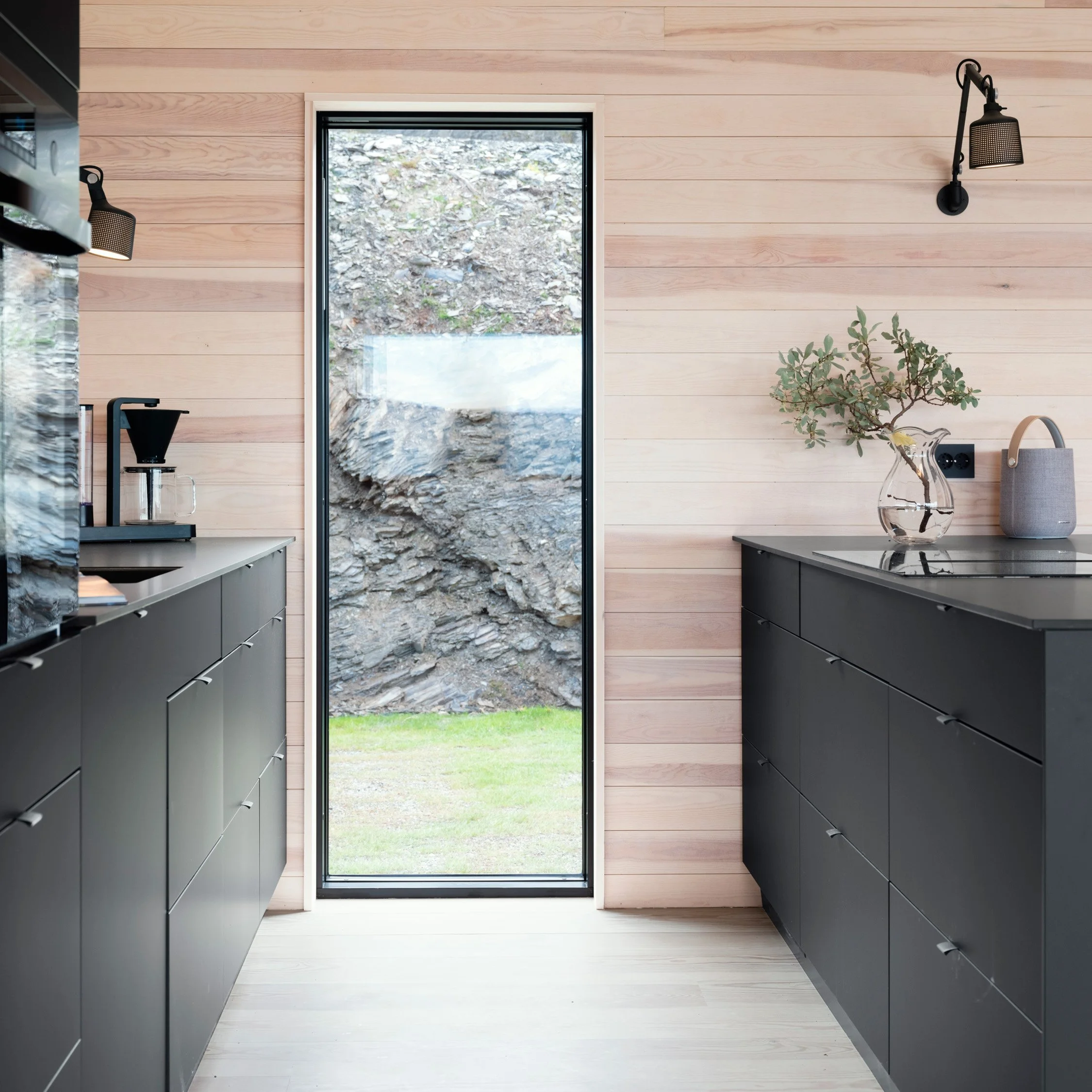 Moderne kjøkken med svarte skap, lyst panel på vegg og et stort vindu som går helt ned til gulvet og som slipper lys og naturen inn.