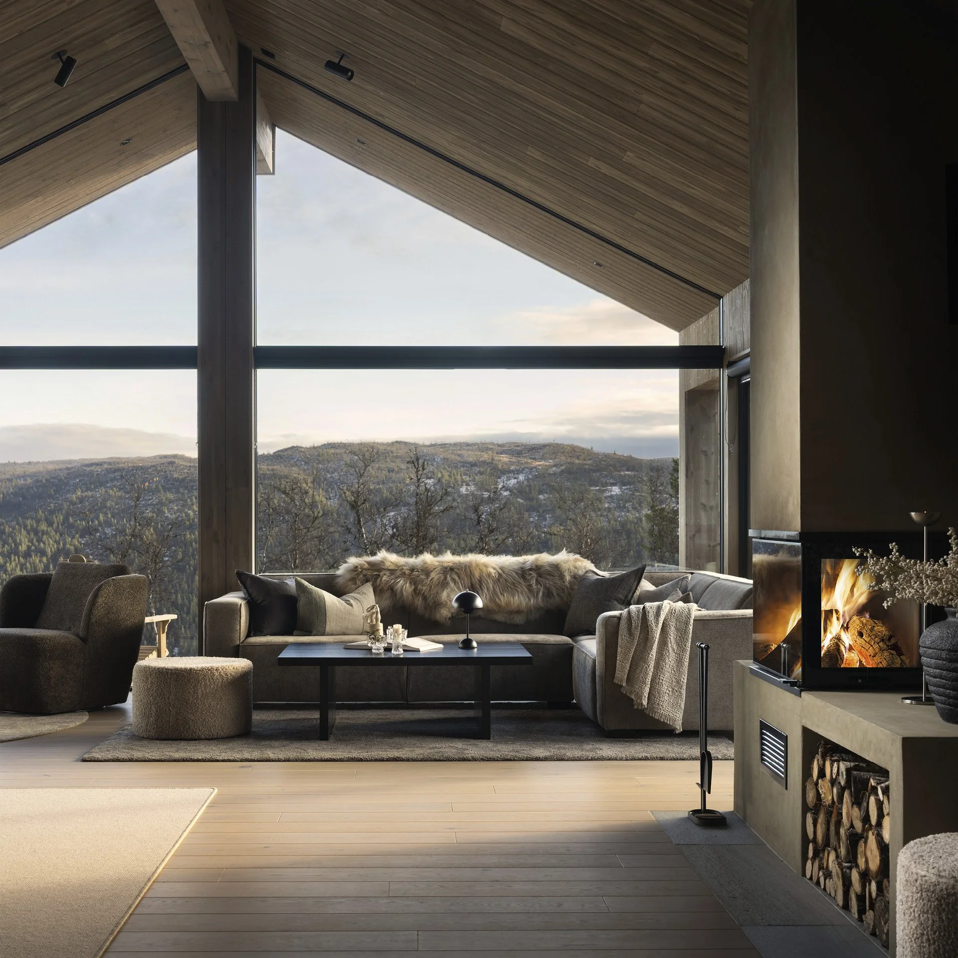 Moderne stue med store vinduer som viser fjelllandskap, med sofa, lenestol, pledd, lampe og peis med fyr i.
