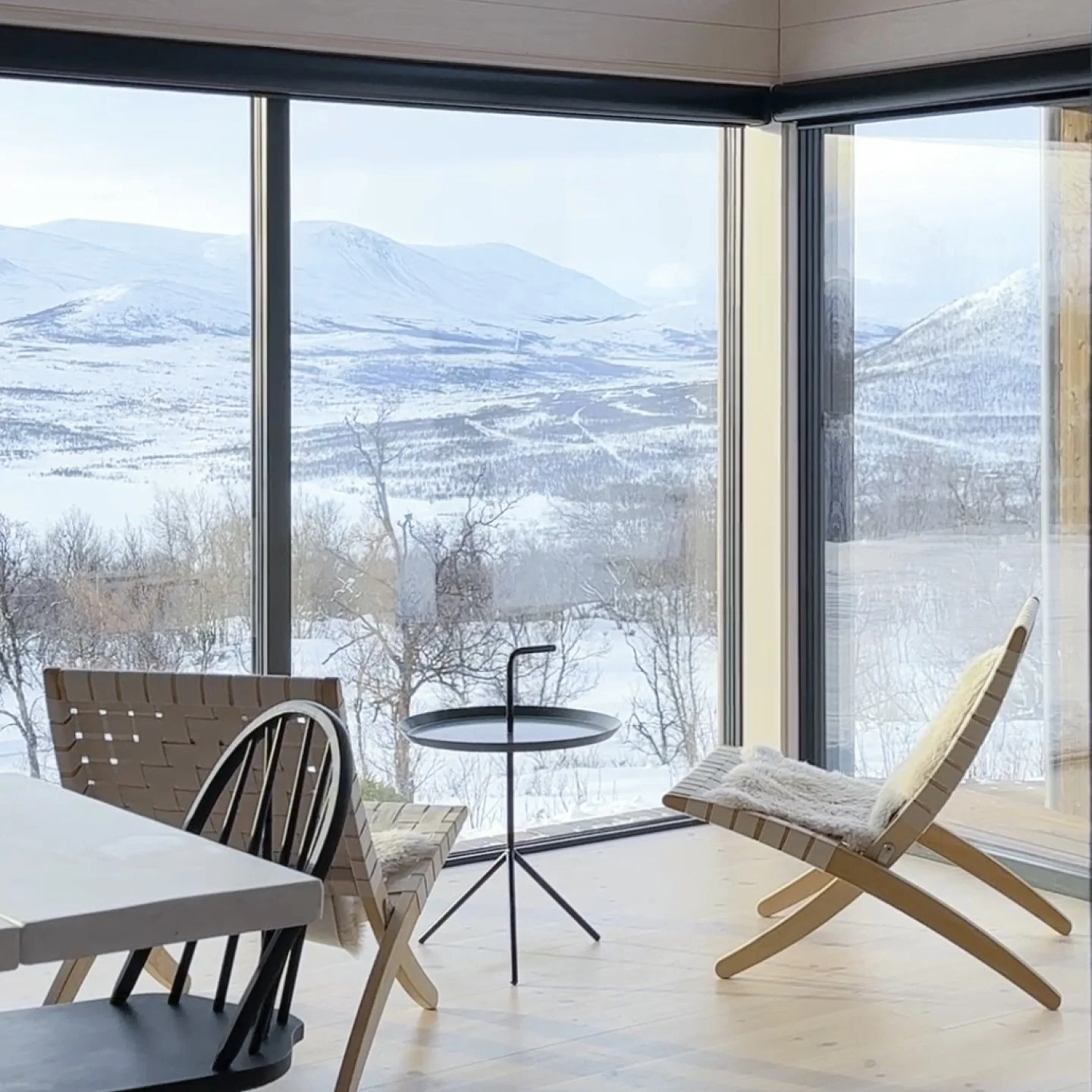 Stue i en hytte, med store hjørnevinduer med utvikt mot et vinterlandskap med fjell og trær i bakgrunnen. Inne står to lenestoler og et lite, sort kaffebord
