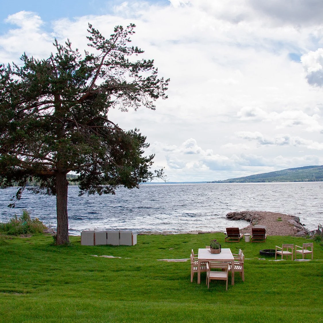 Utsikt over en innsjø med en liten molo. Et stort tre og gressplen med flere sittegrupper i forgrunnen av bildet.