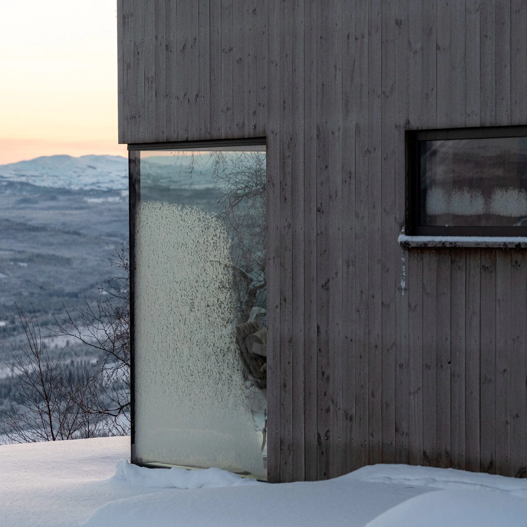 Utvendig bilde av fasade og vindu på hytta. Vinterlandskap med snø og utvikt mot oransje kveldshimmel