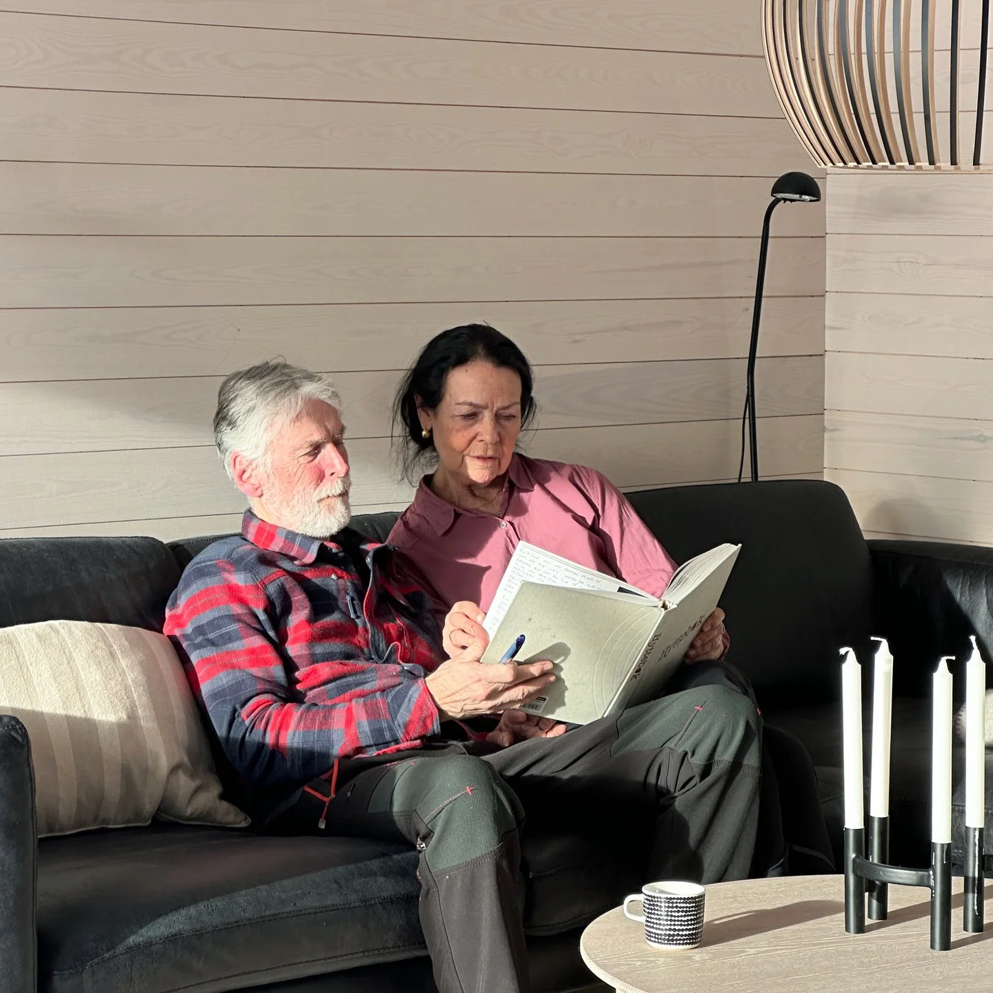 To personer sitter på en svart sofa og leser en bok sammen. Vegger i lyst panel. En kopp står på et bord foran dem, og det er en gulvlampe i bakgrunnen.