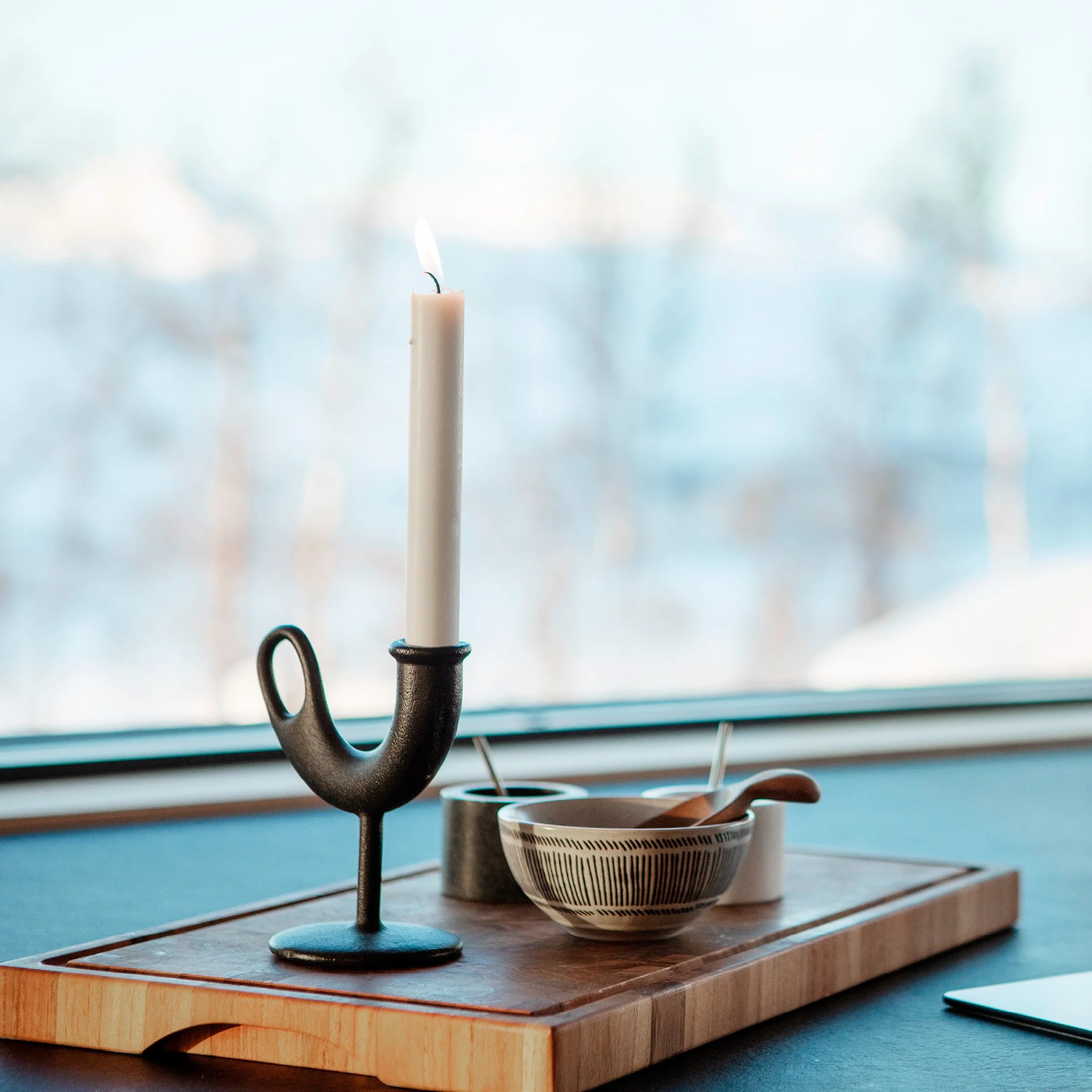 Detaljbilde av sort benkeplate på et kjøkken, med en skjærefjøl, sort moderne lysestake og tre skåler med krydder. I bakgrunnen er det et stort vindu med utsikt mot et vinterlandskap med trær og snødekte fjell.