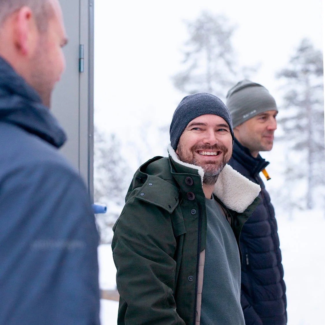 Tre menn står ute i snøvær, smiler og snakker, alle iført jakker og luer.