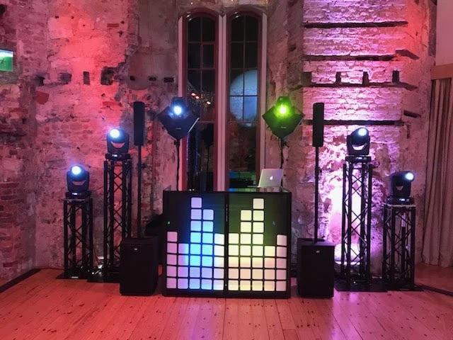 DJ booth setup with colorful LED lights and speakers in a rustic venue with exposed brick walls.