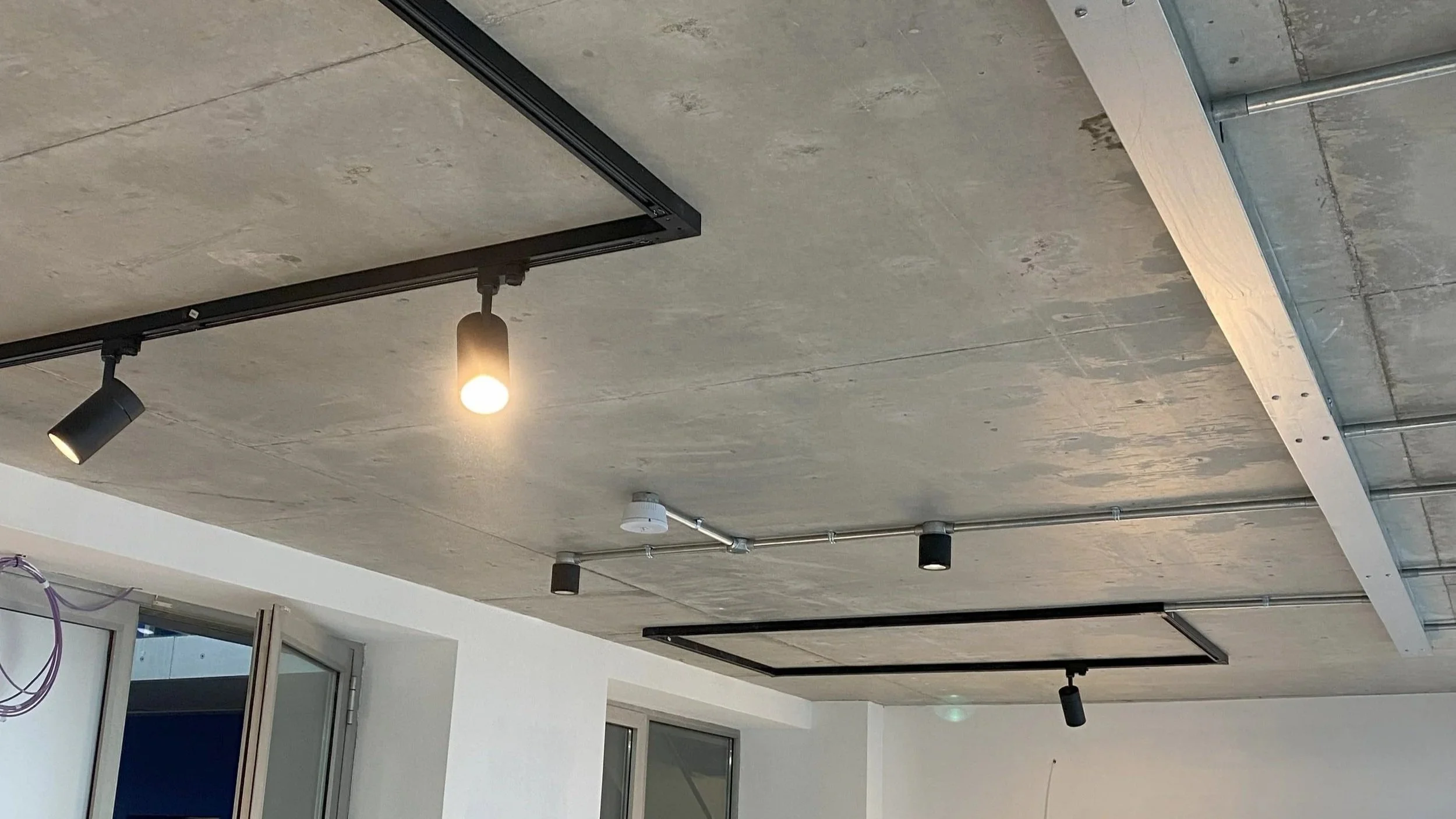 Ceiling with track lighting fixtures, exposed pipes, and an unfinished appearance, in an interior space.
