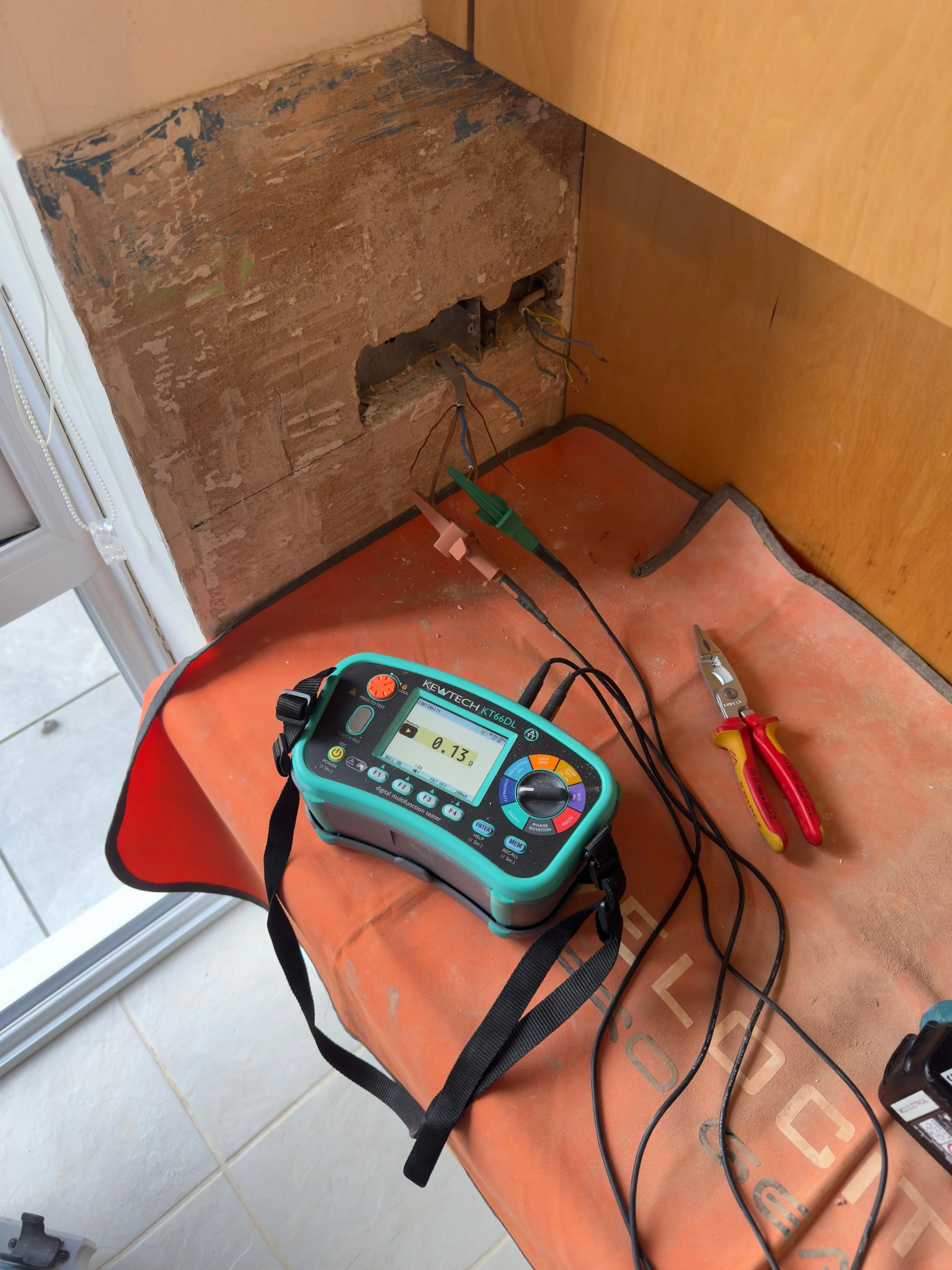 An electrical work setup with wire testing equipment, including a multimeter and wire strippers on an orange work surface. Wires are connected to a wall outlet box, and the background shows a partially removed wall and a wooden cabinet.