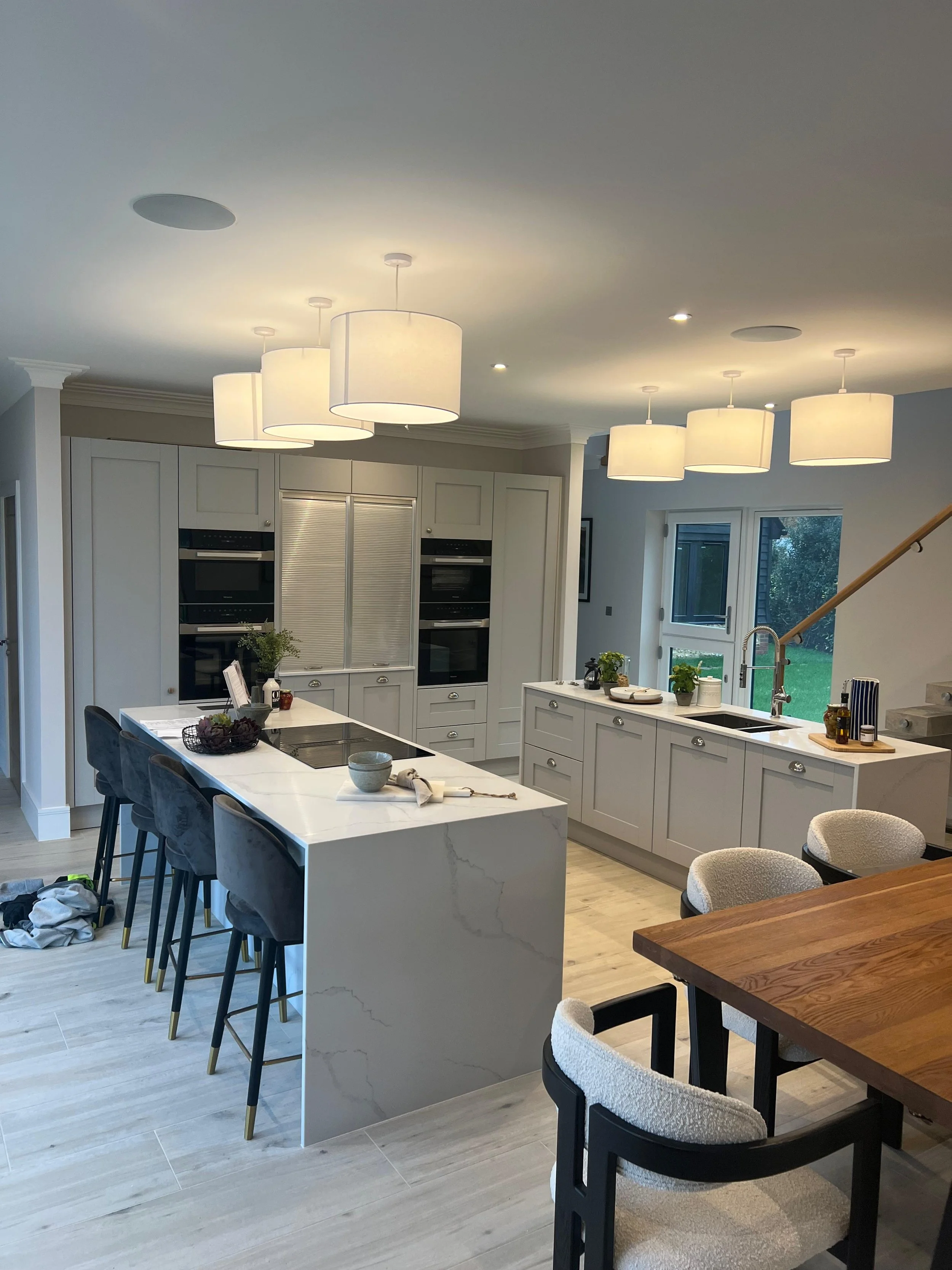Modern kitchen with white cabinets, a central island, bar stools, and a dining table, illuminated by multiple ceiling pendant lights.